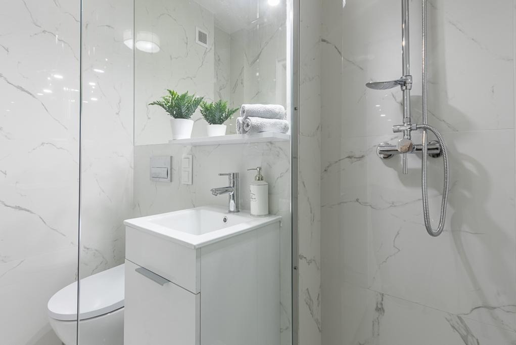 Elegancka, jasna łazienka z marmurowymi płytkami, białą szafką pod umywalką i szklaną kabiną prysznicową. Na półce rośliny w doniczkach i ręczniki.