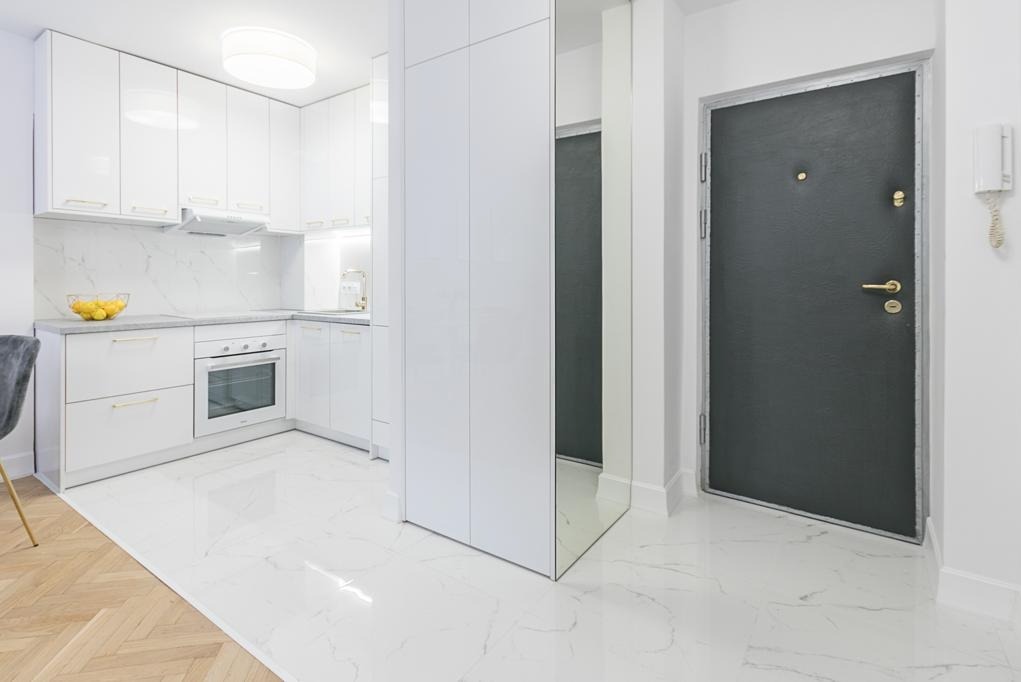 Jasne wnętrze: biała kuchnia z marmurowym blatem, parkiet przechodzi w marmurową podłogę, szara metalowa drzwi wejściowe, minimalizm i nowoczesność.