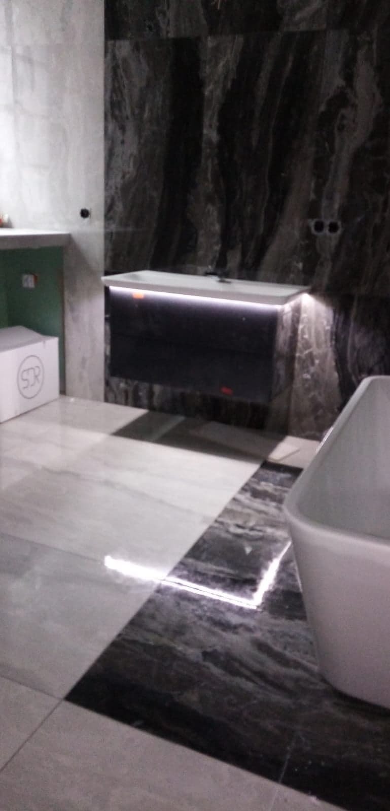 Nowoczesna łazienka z marmurowymi płytkami, podświetlaną szafką pod umywalką i wolnostojącą wanną. Widoczny fragment podłogi i ściany.