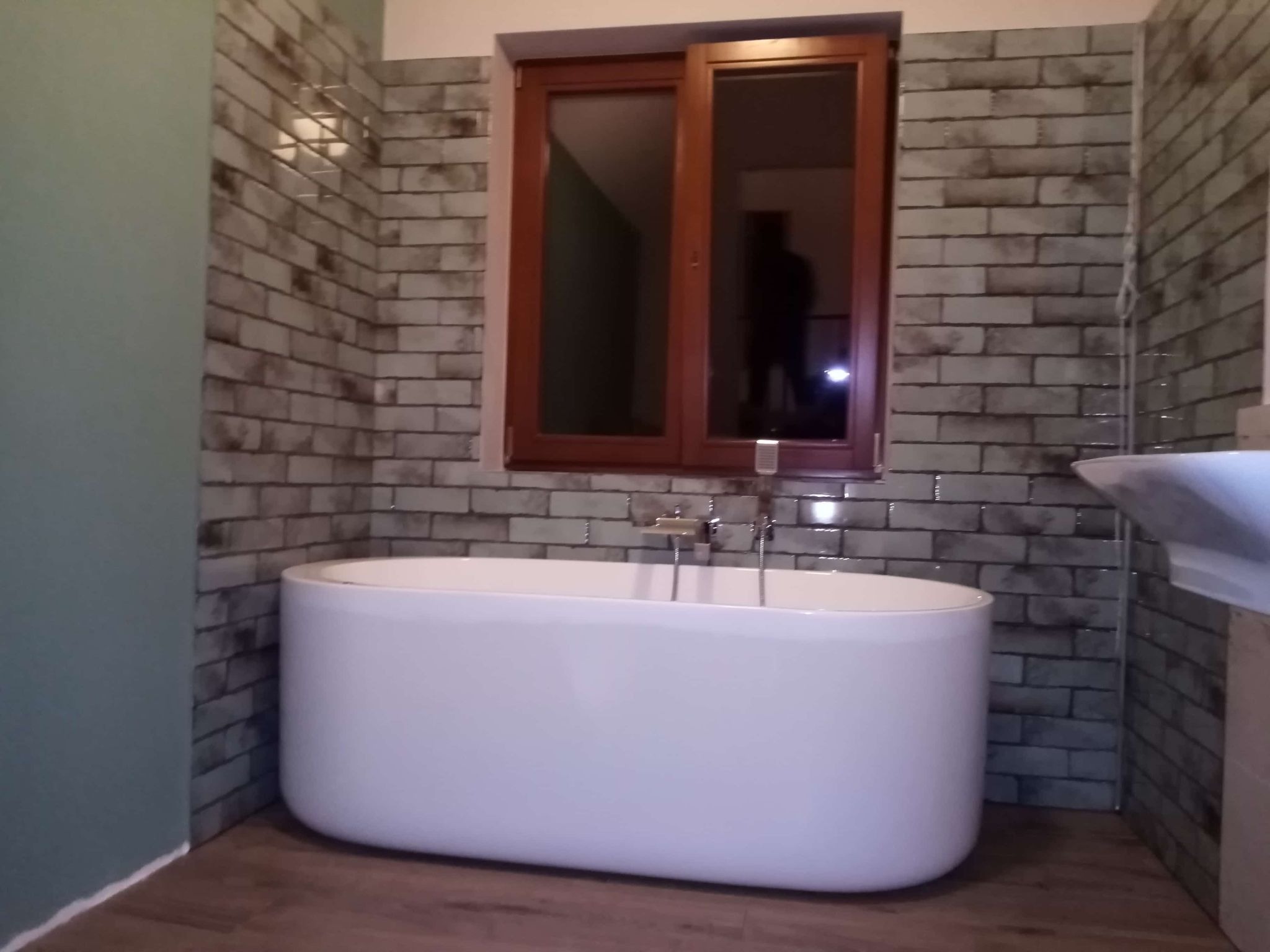 Nowoczesna, biała wanna wolnostojąca na tle ściany wyłożonej szaro-białą cegłą i drewnianego okna. Podłoga z paneli imitujących drewno. Minimalistyczny styl.