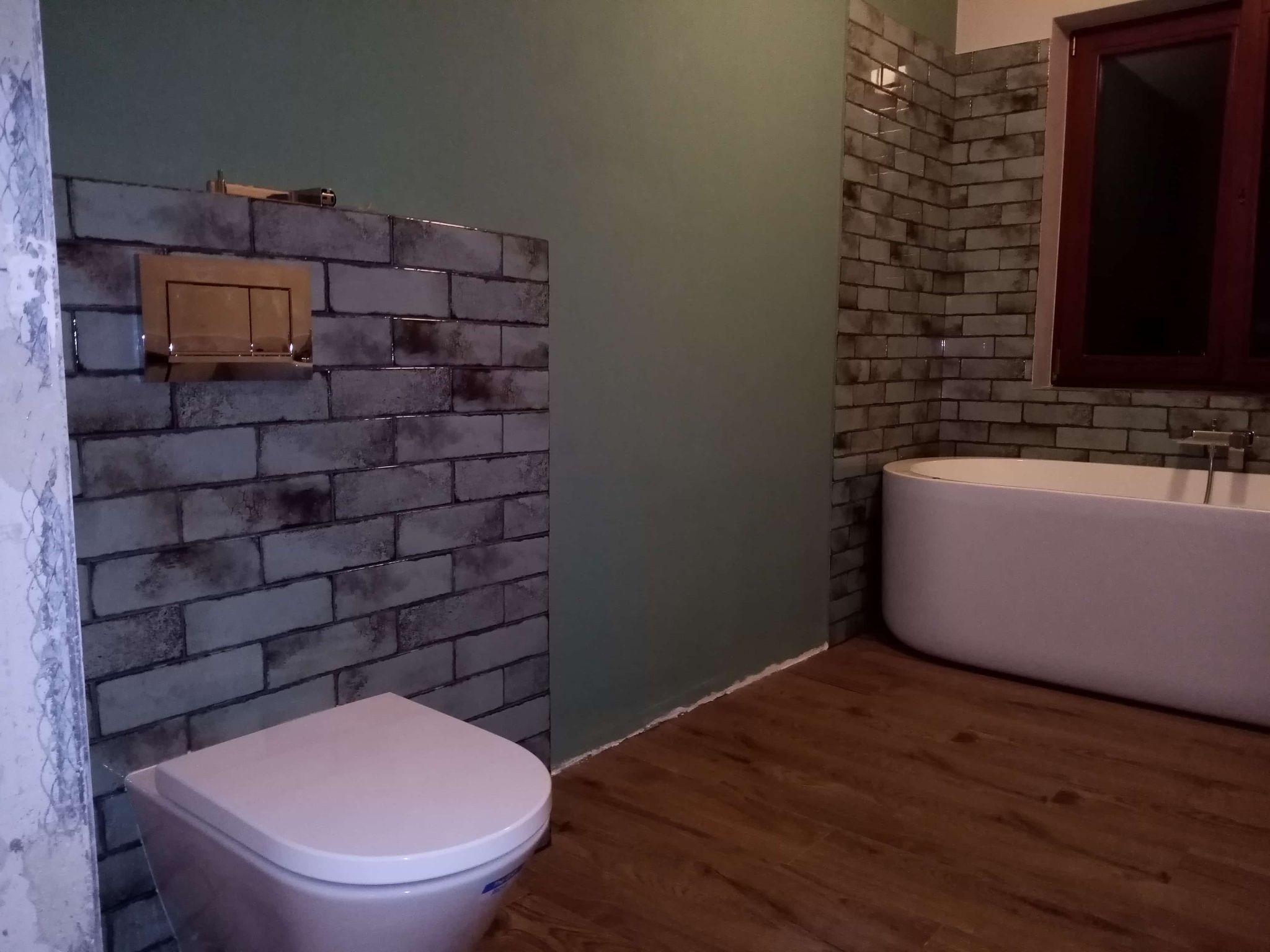 Ukończona łazienka z drewnopodobną podłogą, obudowaną wanną i toaletą. Ściany wyłożone cegiełkowymi płytkami w odcieniach szarości i zieleni. Widoczny fragment okna.