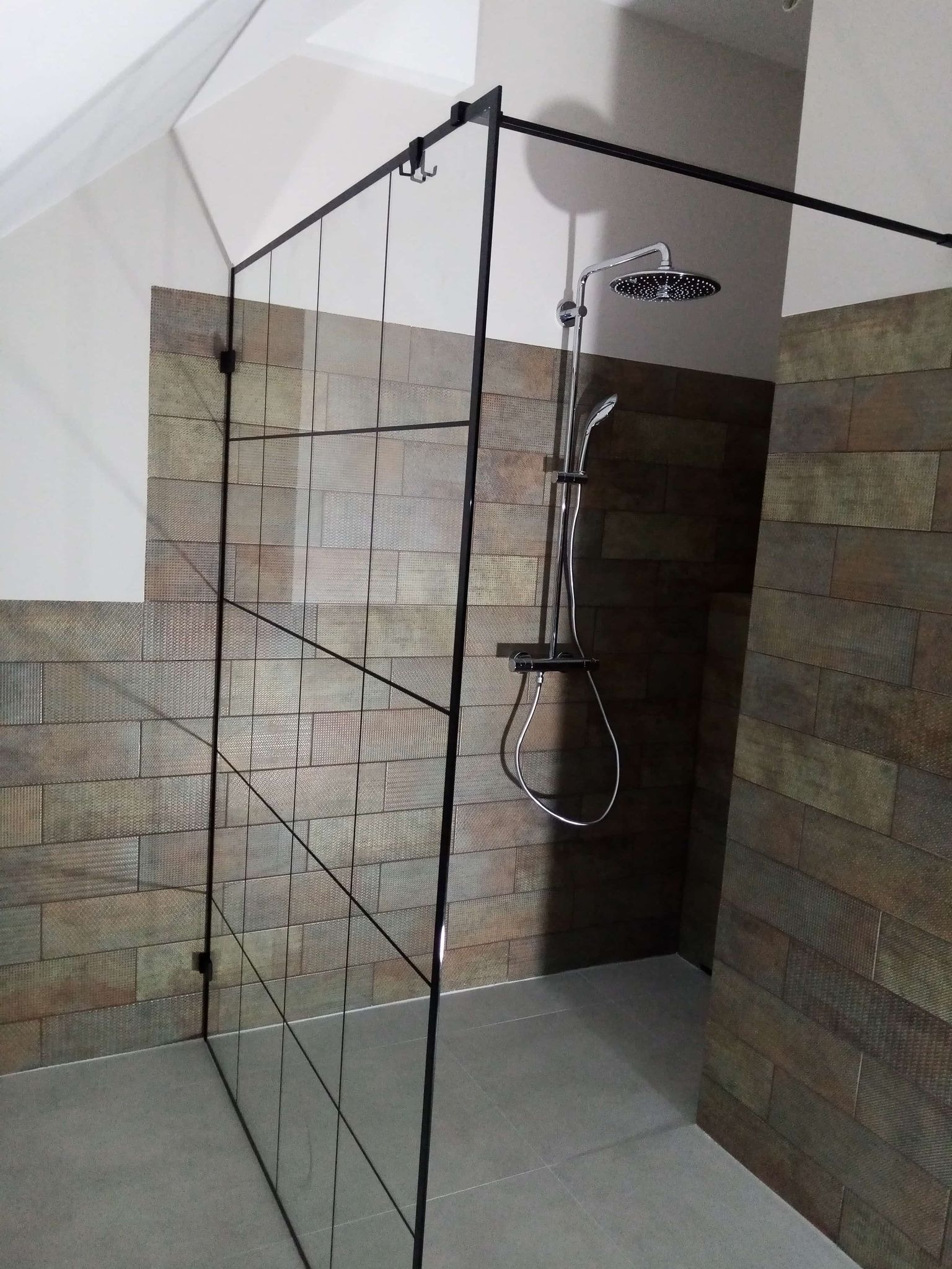 Nowoczesna kabina prysznicowa z czarnymi profilami i deszczownicą, wykończona płytkami imitującymi drewno. Minimalistyczny design, szklana ściana działowa.