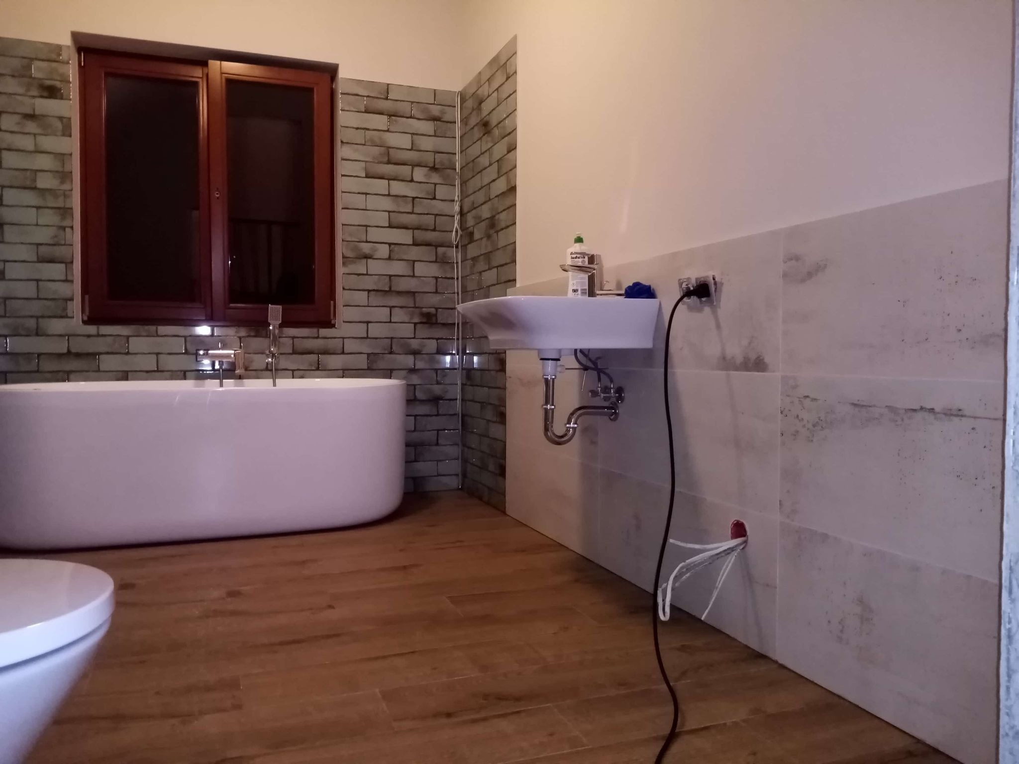 Nowoczesna łazienka z wanną, umywalką i drewnopodobną podłogą. Ściany wyłożone płytkami imitującymi cegłę i marmur. Widoczne gniazdko i przewody.