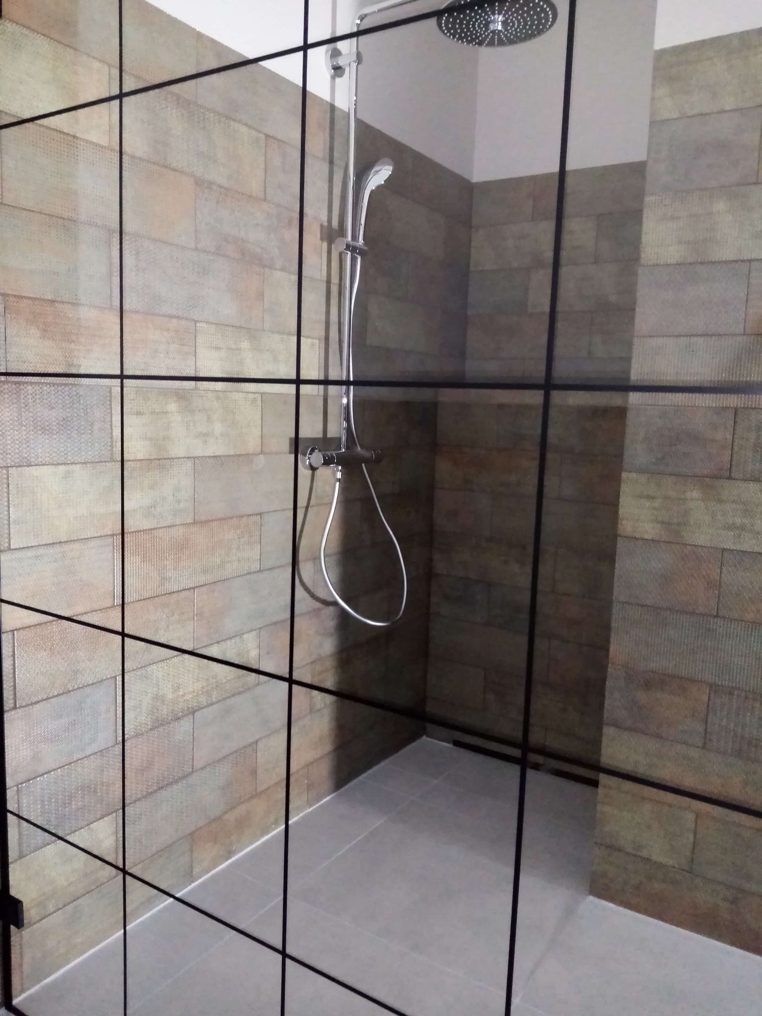 Kabina prysznicowa walk-in z deszczownicą i armaturą w nowoczesnej łazience, wyłożonej płytkami imitującymi drewno. Czarna, geometryczna rama kabiny.