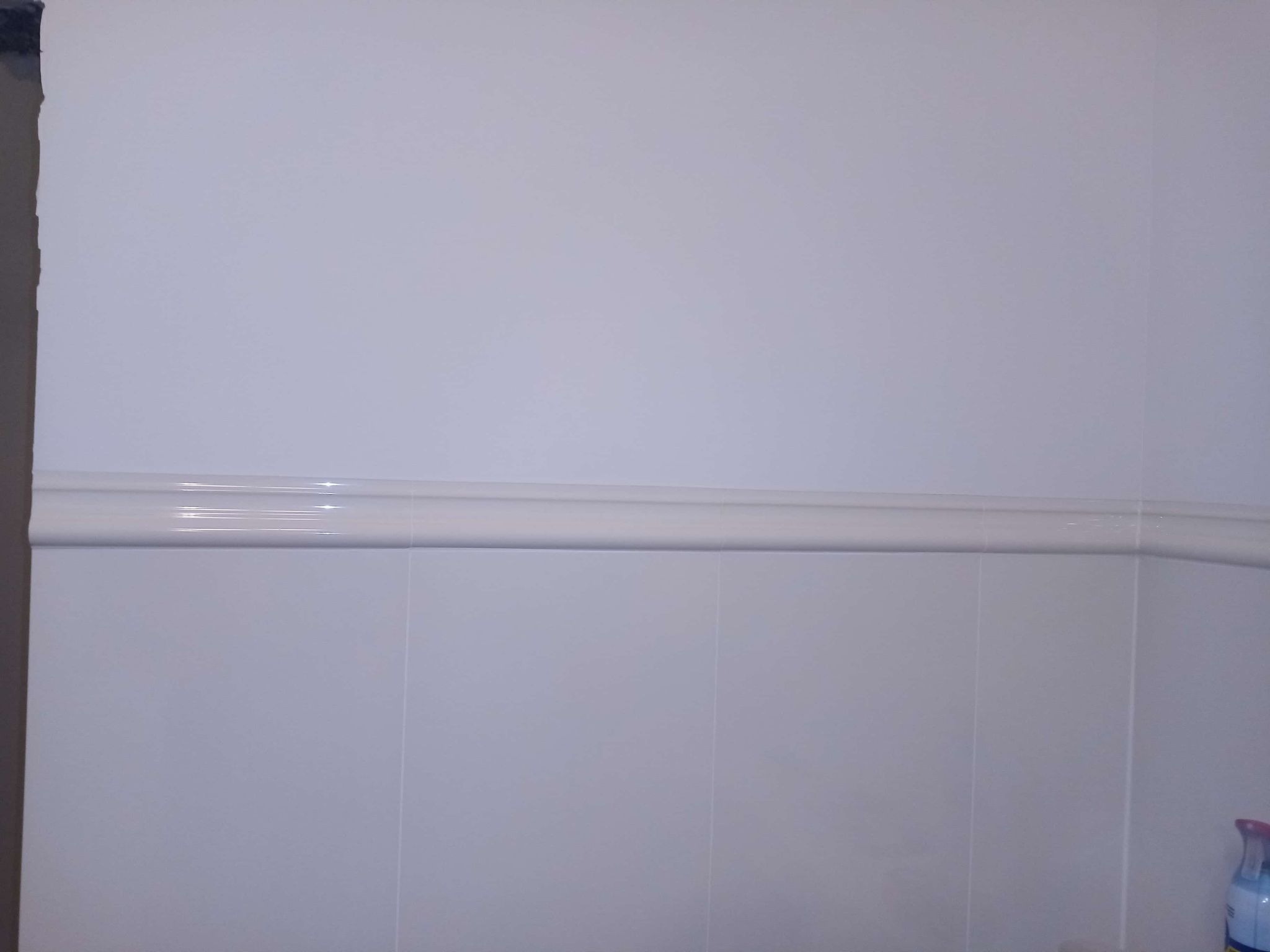 Białe płytki ścienne w łazience, z dekoracyjnym pasem ceramicznym w połowie wysokości. Widoczny fragment ściany i butelka z płynem w rogu.