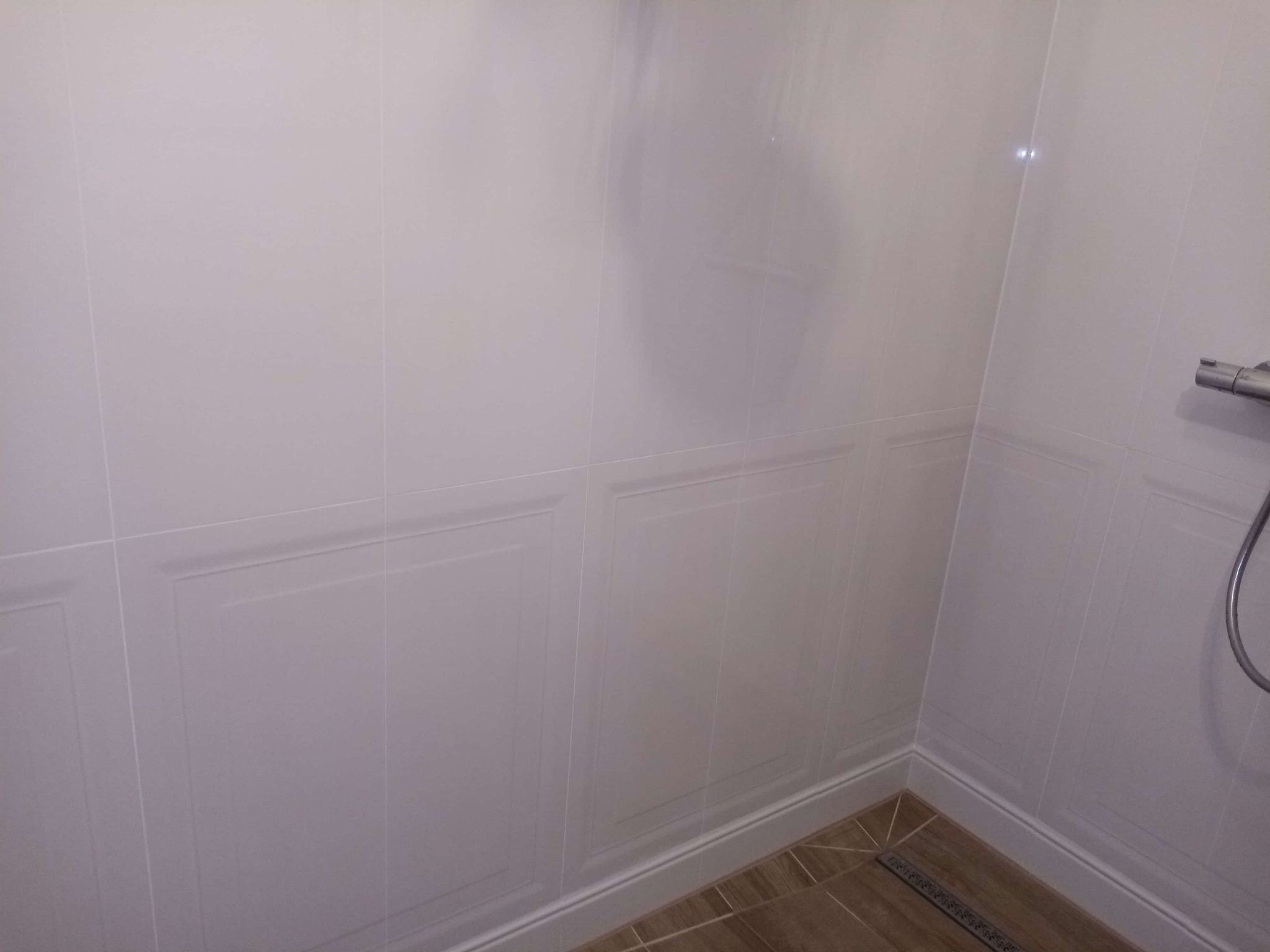 Wykończona łazienka: białe, błyszczące płytki ścienne z subtelnym wzorem paneli, brązowa podłoga z odpływem liniowym. Widoczny fragment baterii prysznicowej.