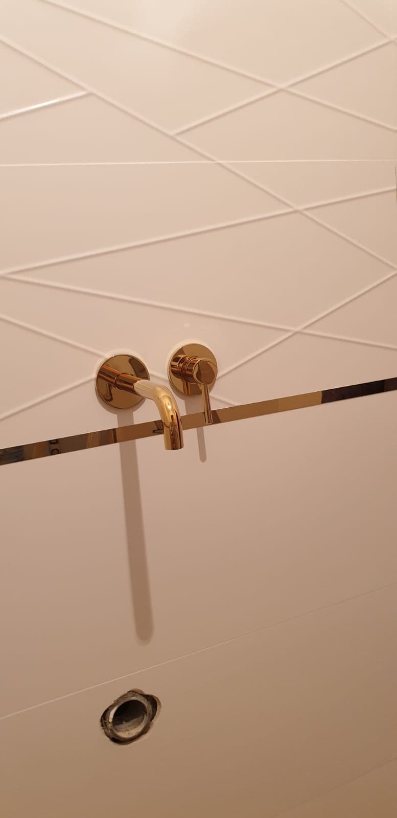 Białe płytki z geometrycznym wzorem i złotą armaturą łazienkową, widoczne otwór odpływowy. Minimalistyczny design z akcentem luksusu.