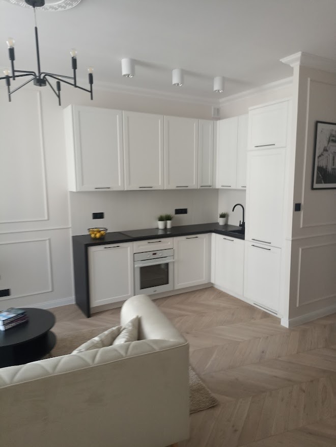 Jasna kuchnia w stylu klasycznym: białe szafki z czarnym blatem, drewniana podłoga w jodełkę, nowoczesne oświetlenie. Widok z salonu, sofa w kremowym odcieniu.