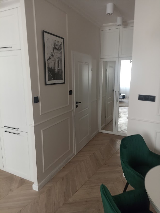 Eleganckie wykończenie wnętrza: białe szafy w zabudowie, parkiet w jodełkę, drzwi z frezowanymi panelami i ciemnozielone krzesła przy okrągłym stole.