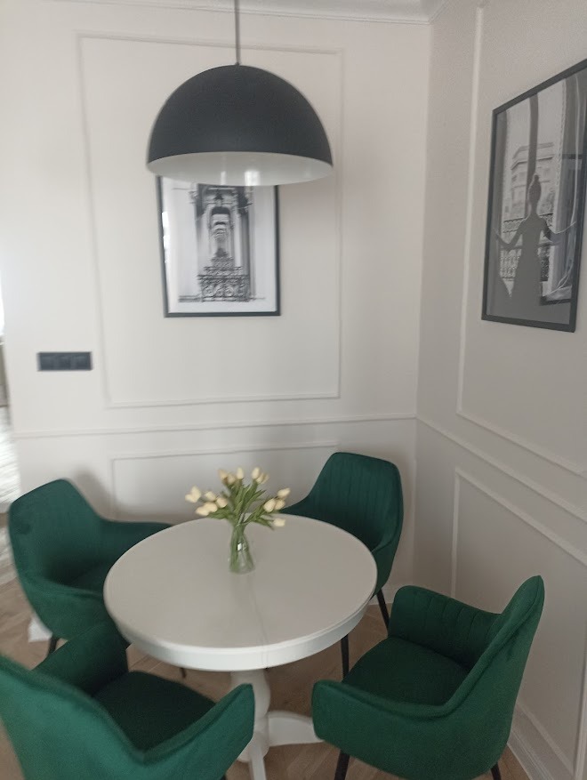 Elegancka jadalnia: okrągły biały stół, zielone krzesła, czarna lampa wisząca i obrazy w ramach na ścianie z dekoracyjną sztukaterią. Nowoczesny styl, jasne wnętrze.