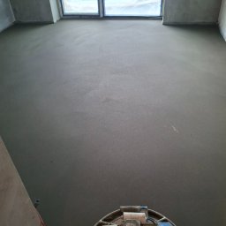 Piotr Komperda wylewki maszynowe - Gładka, szara posadzka po wylaniu, zacierana maszynowo. Widoczne okno z folią ochronną. Perspektywa od dołu, z maszyną do zacierania w kadrze.