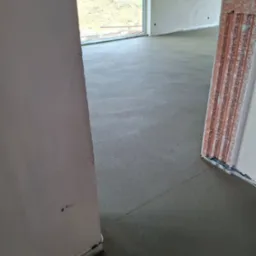 Świeżo wylana, gładka posadzka w jasnym wnętrzu, widok z poziomu podłogi, z fragmentami ścian i okna w tle. Wykończona powierzchnia gotowa do dalszych prac.