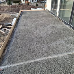 Piotr Komperda wylewki maszynowe - Świeżo wylana, szara posadzka na tarasie domu jednorodzinnego, z widocznym drewnianym szalunkiem i oknami w tle. Gotowa powierzchnia pod wykończenie.