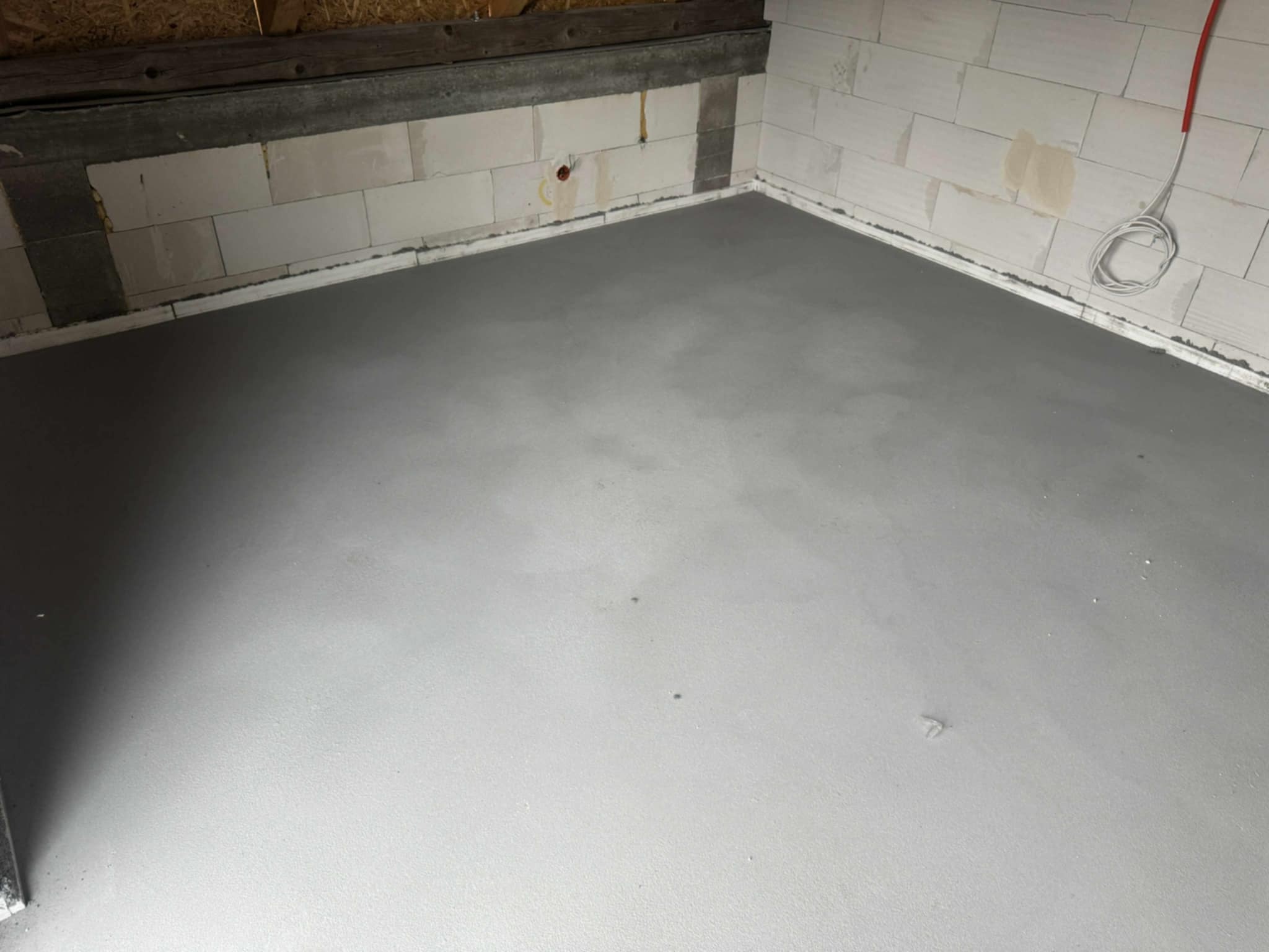 Świeżo wylana, gładka, szara posadzka jastrychowa w pomieszczeniu z białymi ścianami z betonu komórkowego i widoczną instalacją elektryczną.
