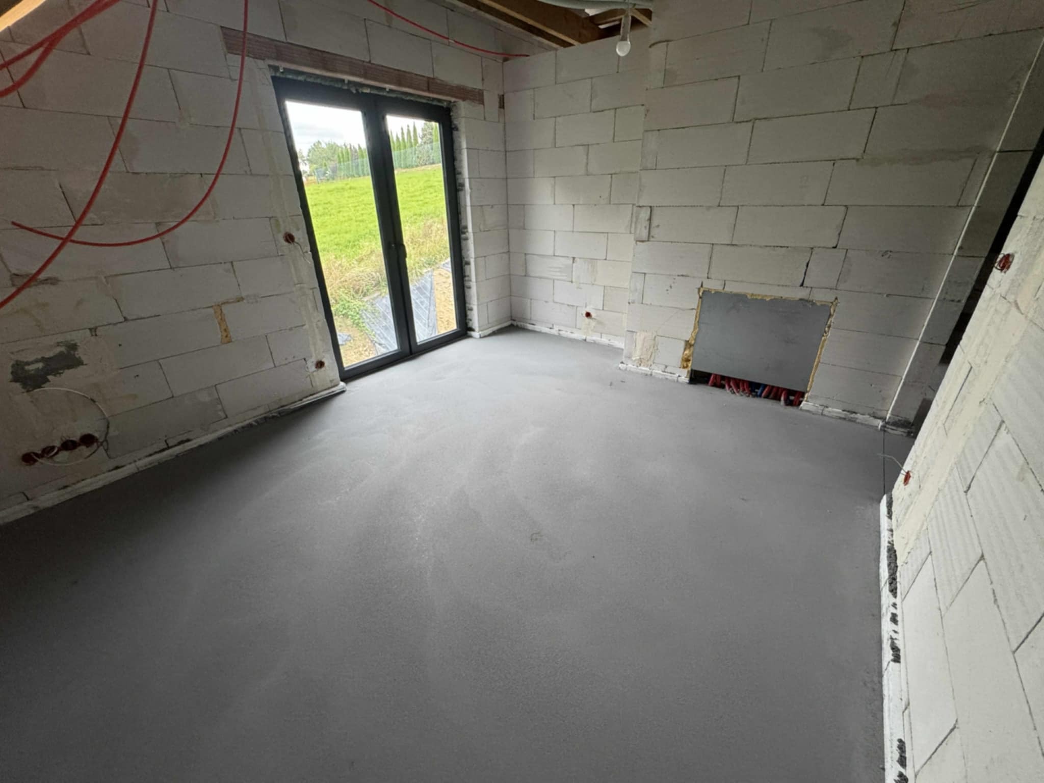 Szara, gładka wylewka w surowym wnętrzu z białymi bloczkami, widok na okno i instalacje elektryczne. Minimalistyczny styl wykończenia podłogi.