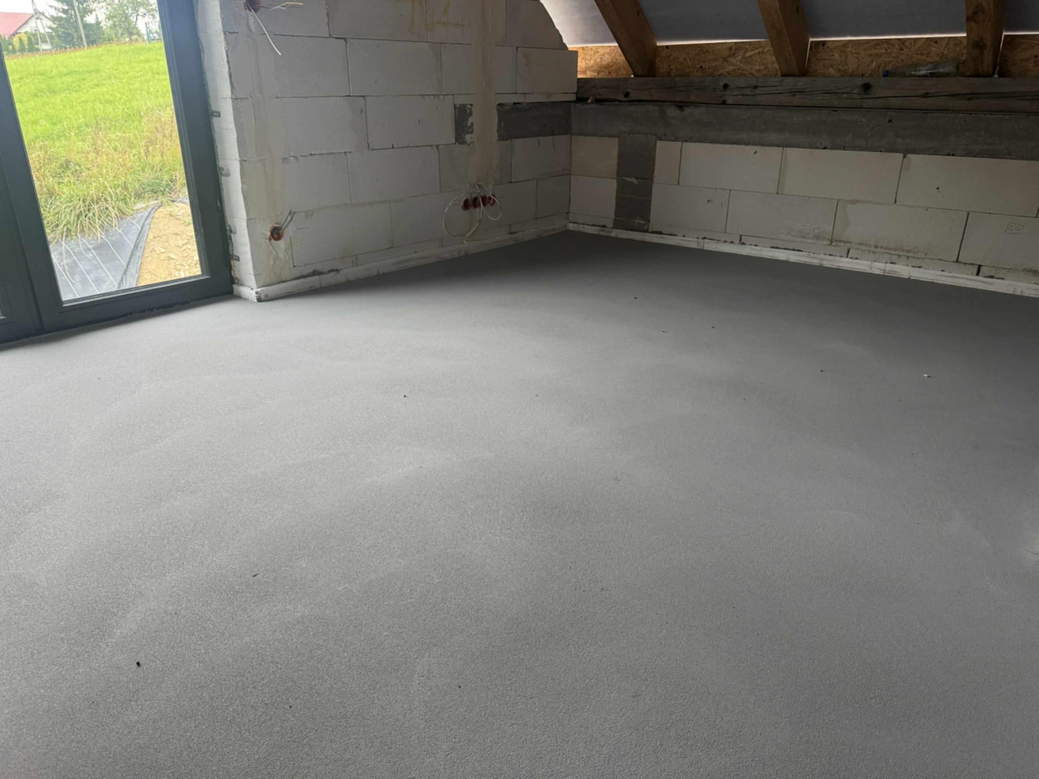 Gładka, szara posadzka w surowym wnętrzu z białymi bloczkami i widoczną konstrukcją dachu. Okno z widokiem na zieleń. Widoczne kable elektryczne.