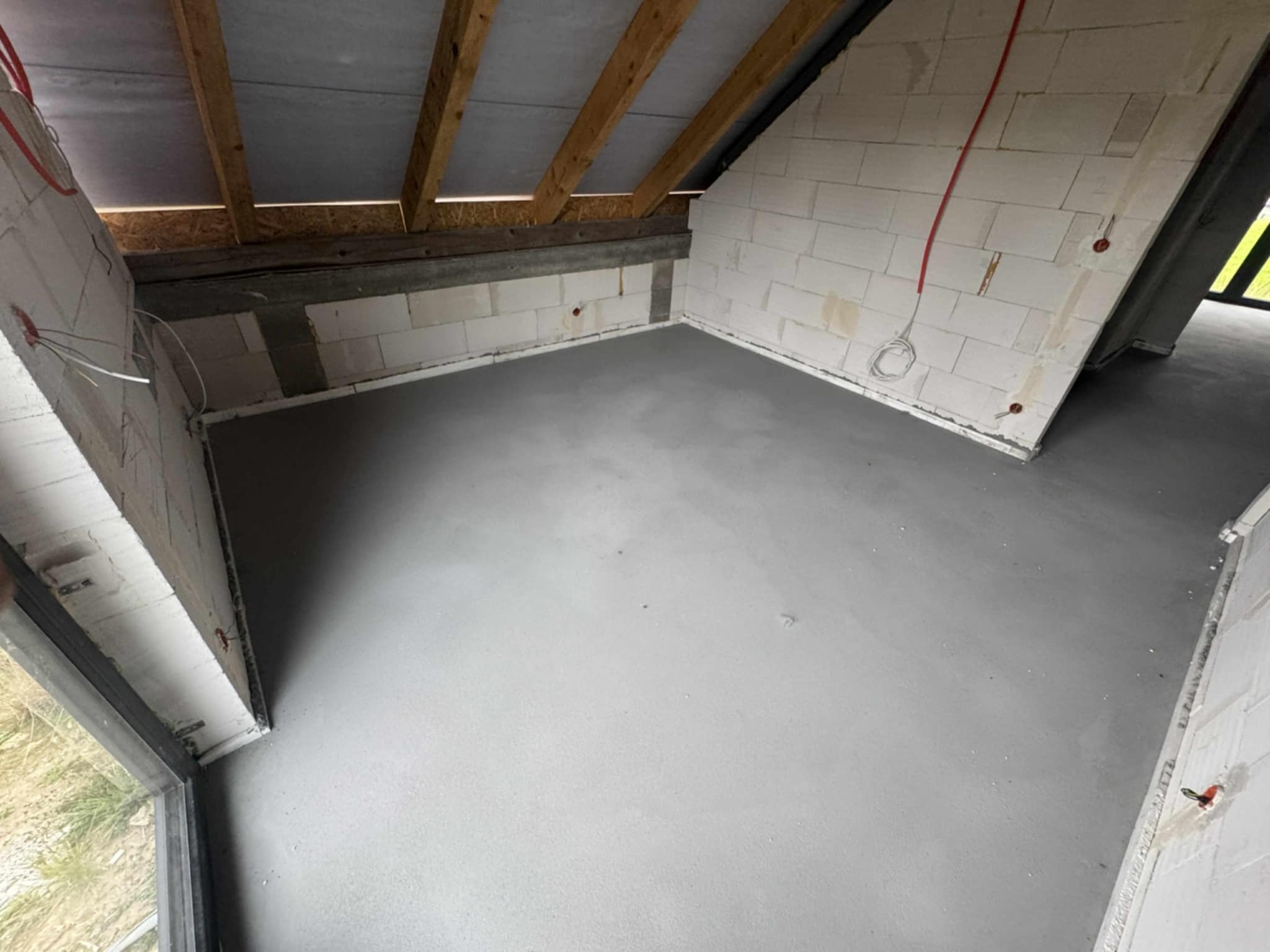 Szara, gładka posadzka w surowym wnętrzu poddasza z oknem i fragmentami instalacji elektrycznej na ścianach z betonu komórkowego. Widoczne drewniane krokwie.