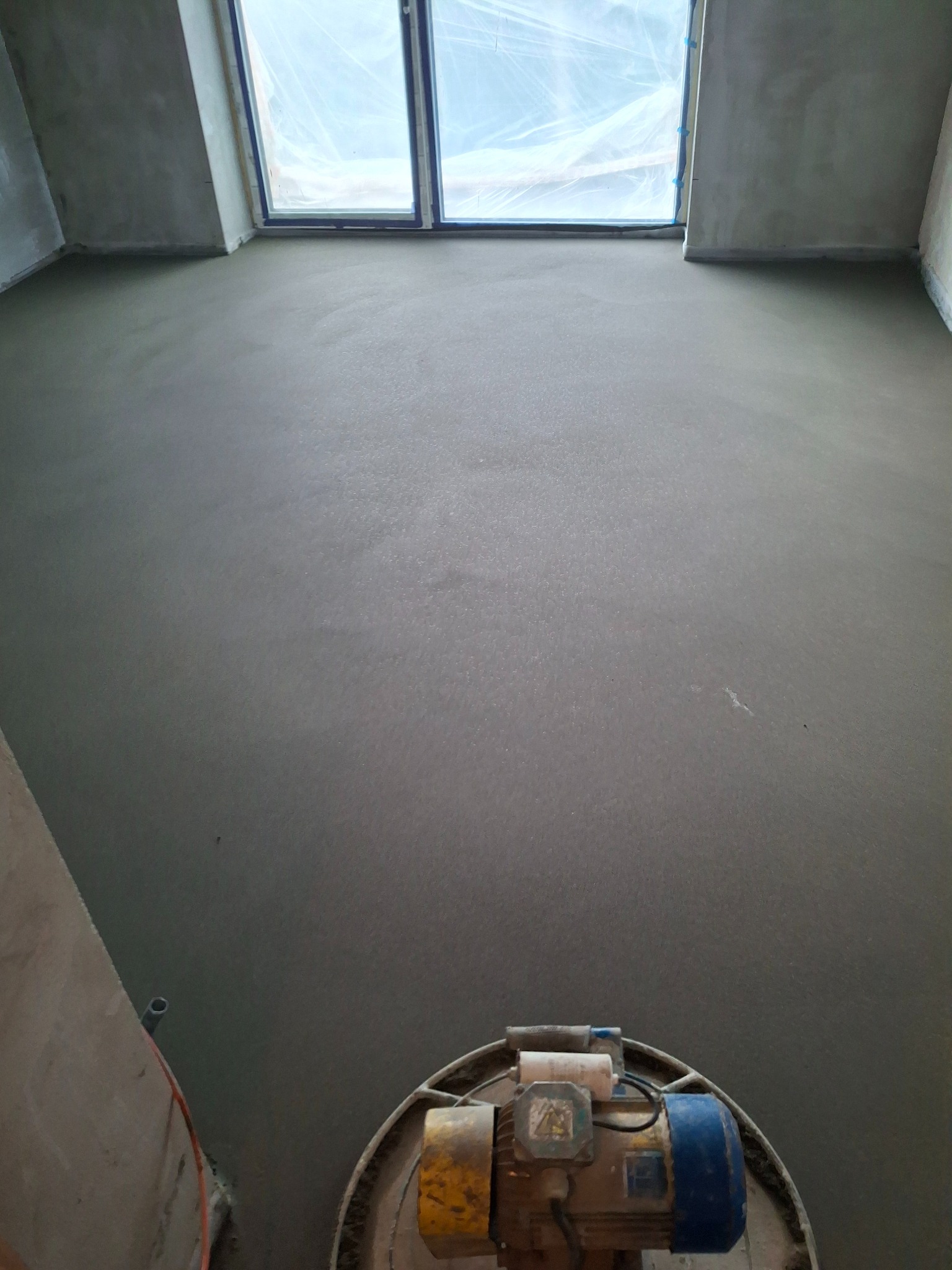 Gładka, szara posadzka po wylaniu, zacierana maszynowo. Widoczne okno z folią ochronną. Perspektywa od dołu, z maszyną do zacierania w kadrze.