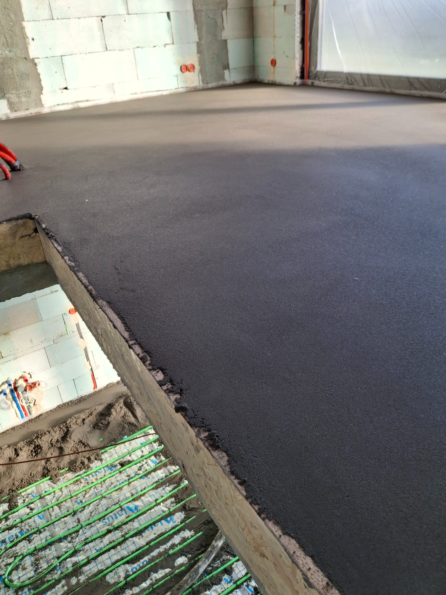 Świeżo wylana, ciemna posadzka w surowym wnętrzu, z widocznym systemem ogrzewania podłogowego w wykopie. Surowe ściany z białego bloczka.