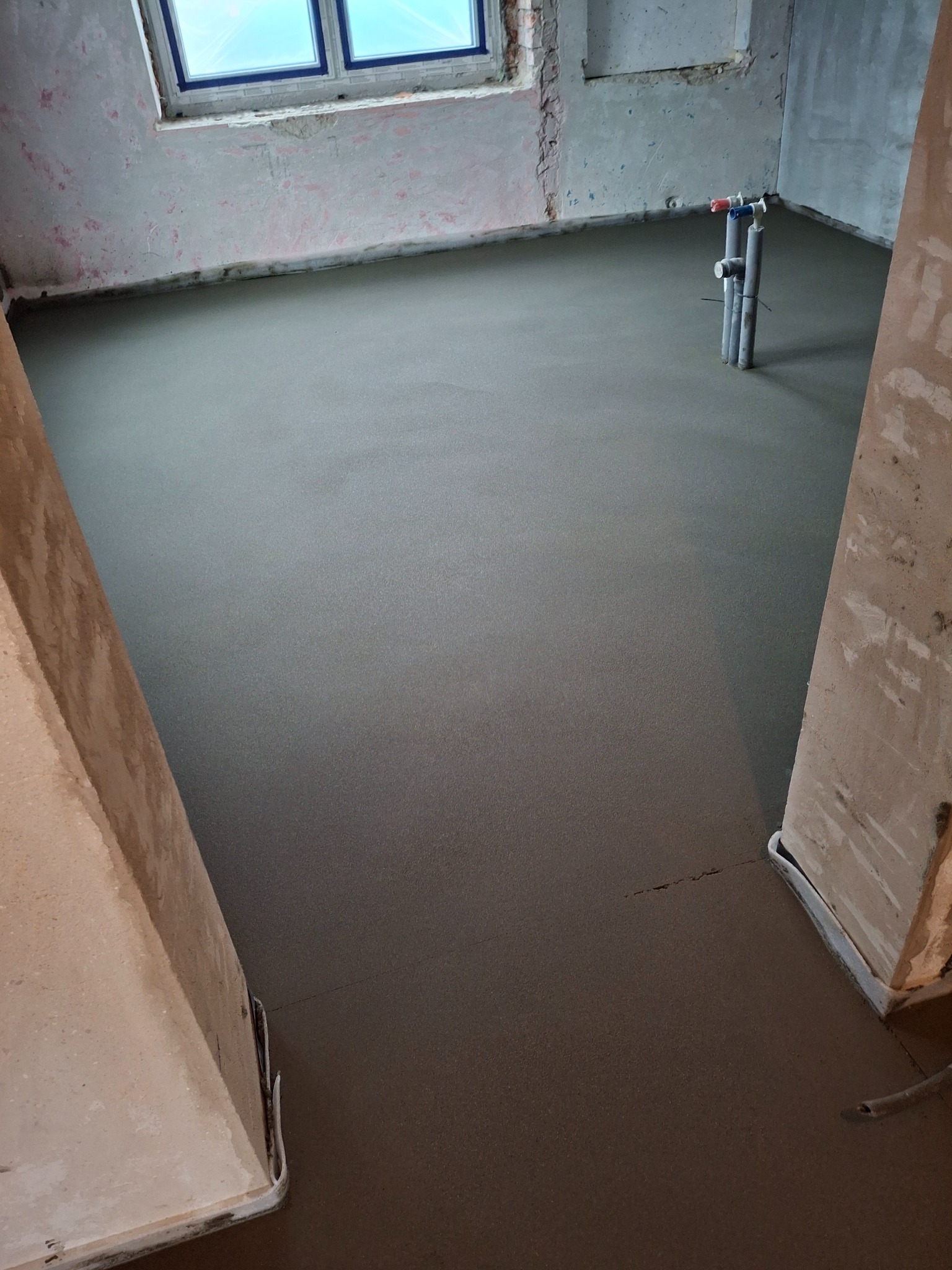 Świeżo wylana, gładka posadzka w surowym wnętrzu z widocznymi rurami instalacyjnymi i fragmentem okna. Minimalistyczny, szary odcień posadzki.