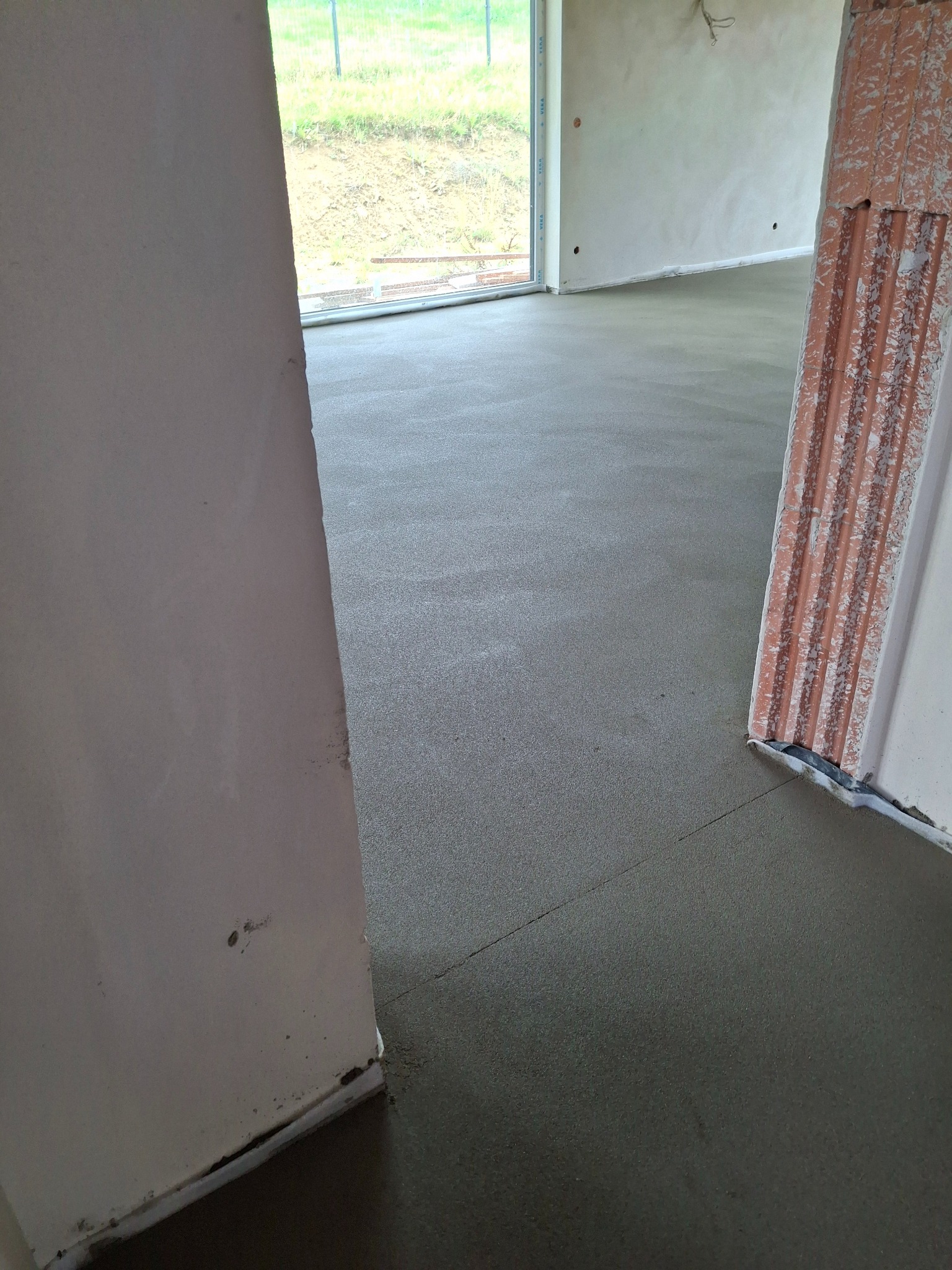 Świeżo wylana, gładka posadzka w jasnym wnętrzu, widok z poziomu podłogi, z fragmentami ścian i okna w tle. Wykończona powierzchnia gotowa do dalszych prac.