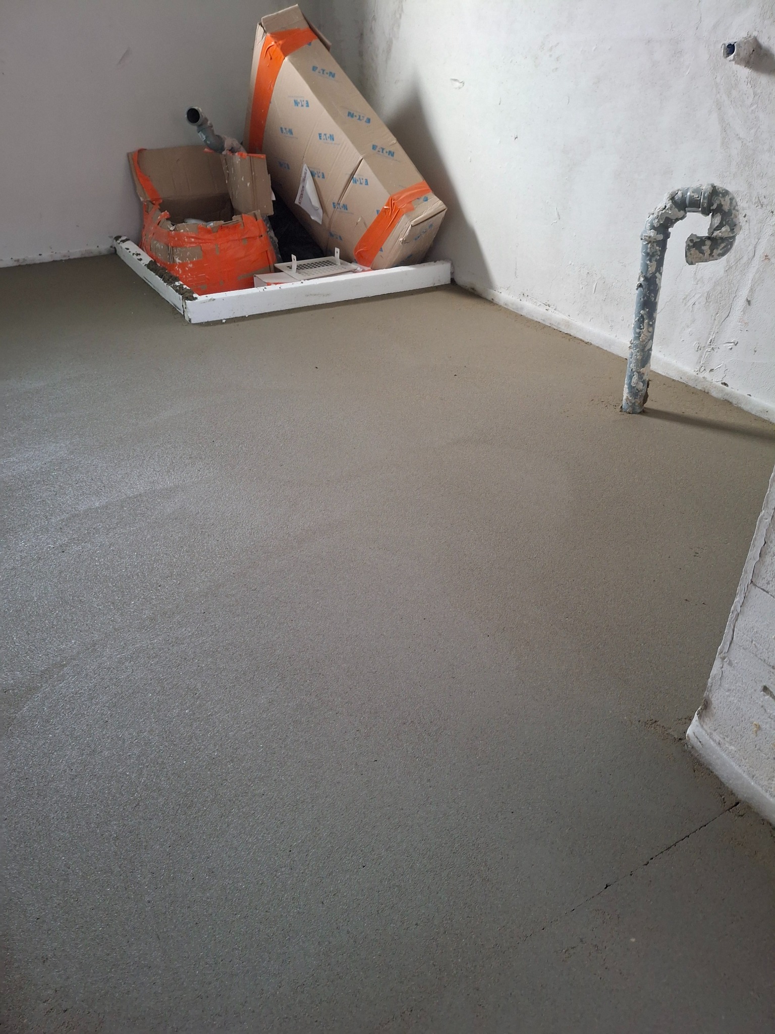 Świeżo wylana, gładka posadzka w pomieszczeniu. Widoczne kartony i rura przy ścianie. Jasnoszara powierzchnia, delikatna tekstura. Prace wykończeniowe w toku.