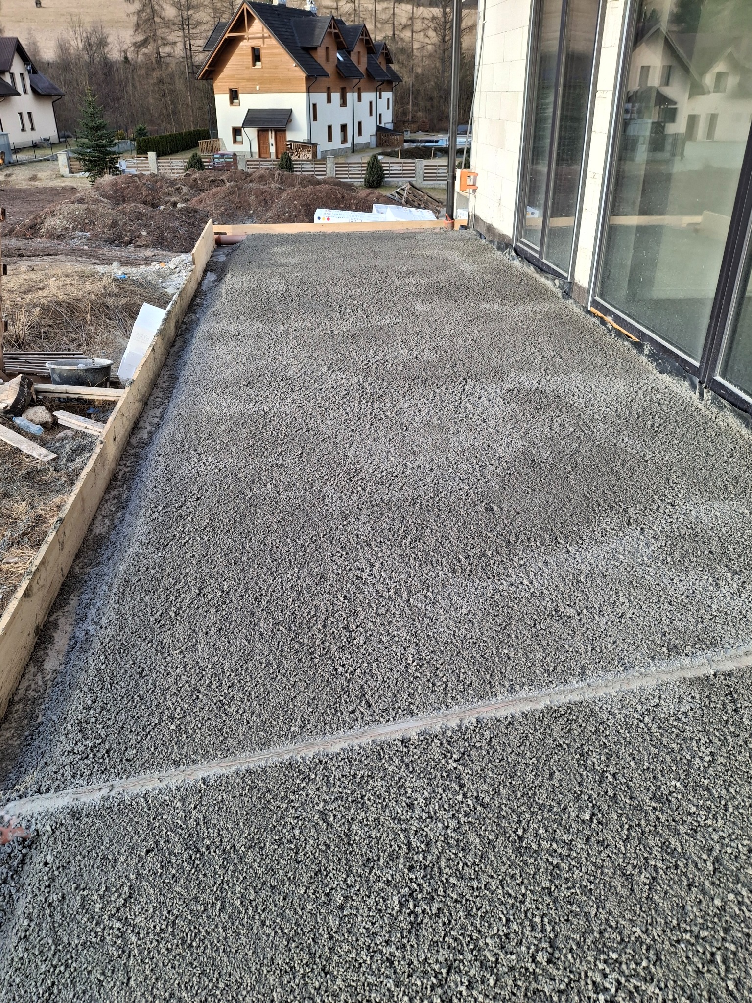 Świeżo wylana, szara posadzka na tarasie domu jednorodzinnego, z widocznym drewnianym szalunkiem i oknami w tle. Gotowa powierzchnia pod wykończenie.