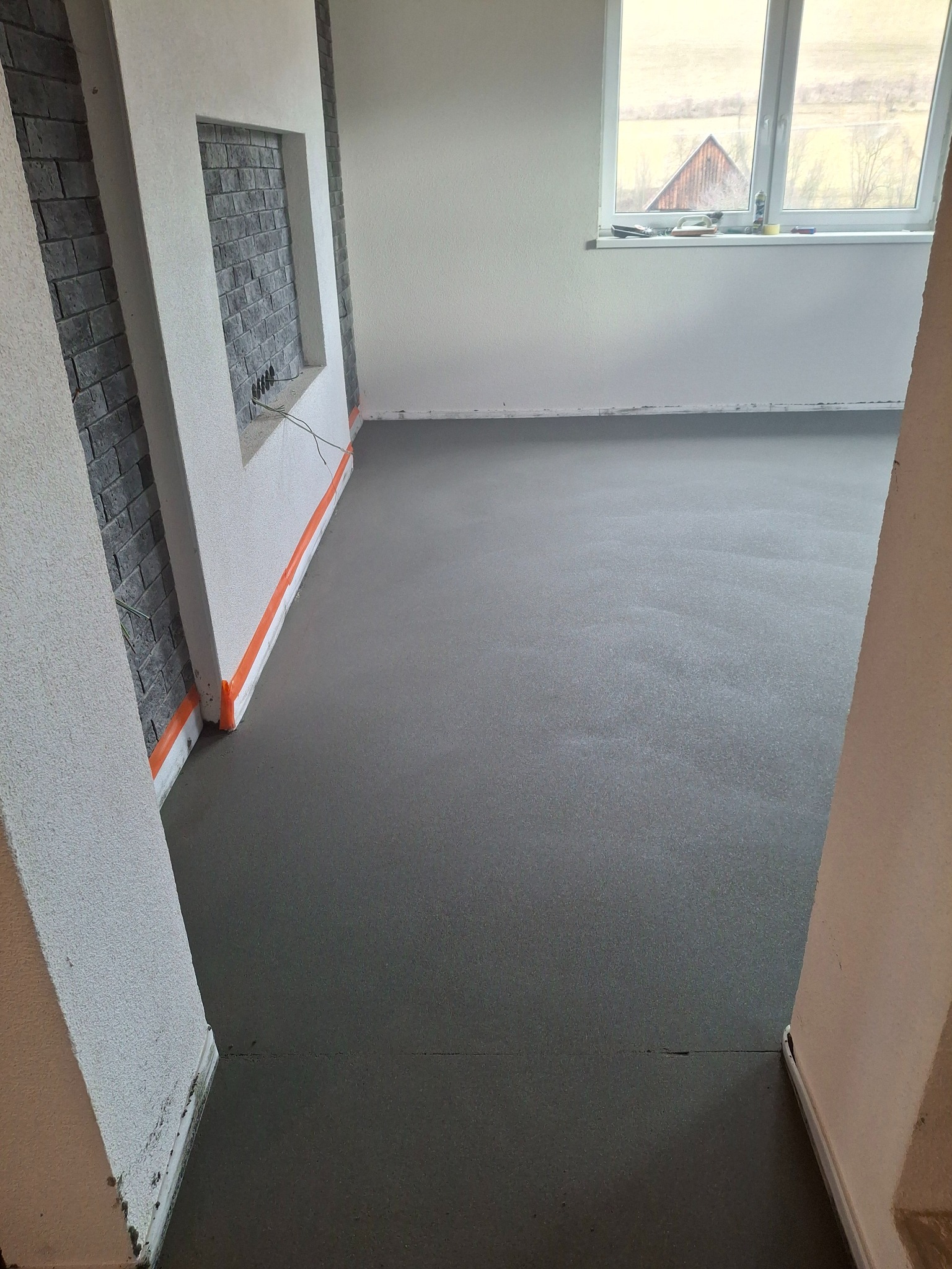 Świeżo wylana, szara posadzka jastrychowa w jasnym pomieszczeniu z oknem. Widoczne taśmy zabezpieczające ściany i fragment dekoracyjnej ściany z cegły.