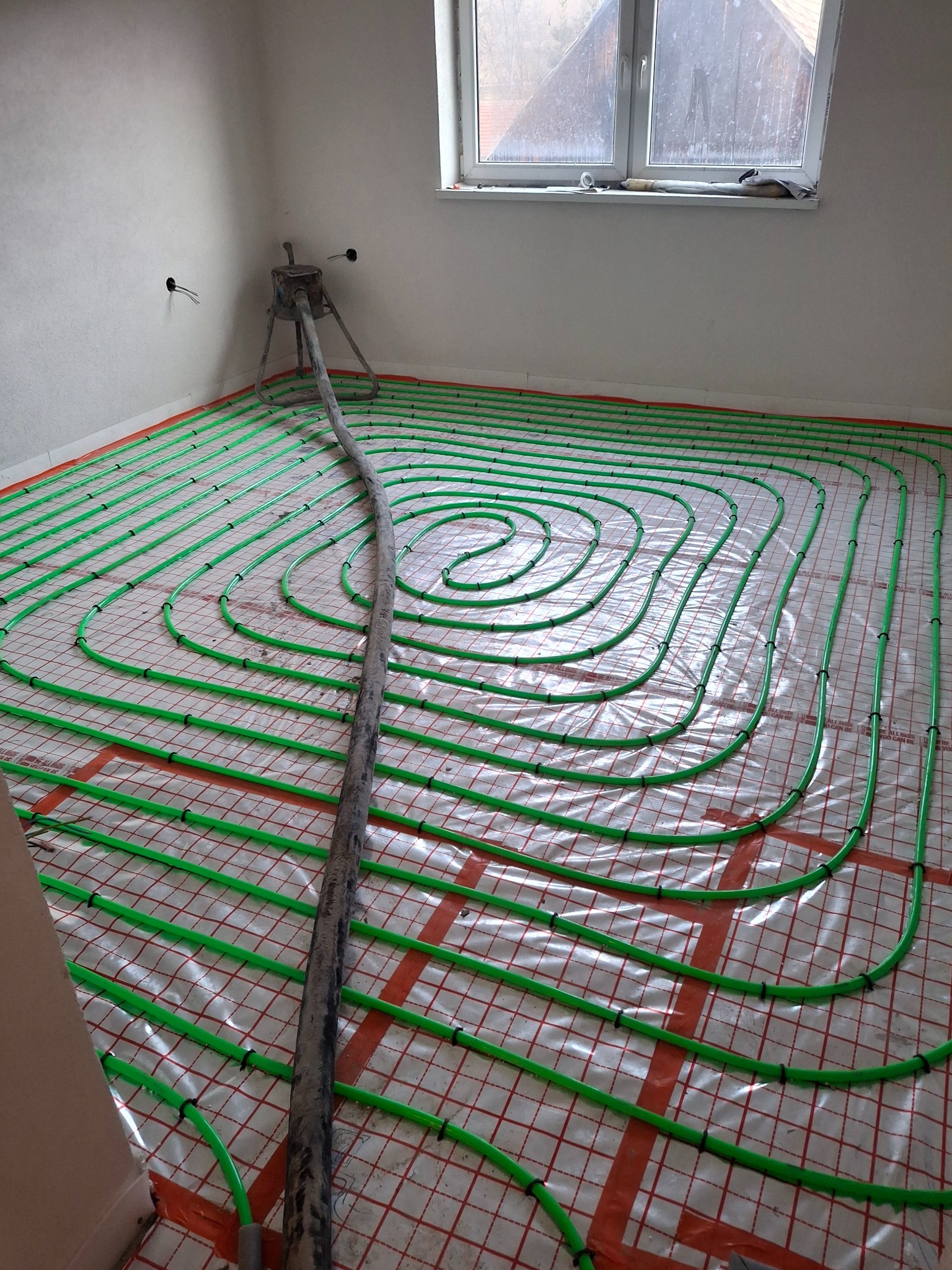 Ogrzewanie podłogowe: zielone rurki na folii z kratką, podłączone do pompy. Widok na okno i surowe ściany. Etap instalacji przed wylaniem posadzki w nowym domu.