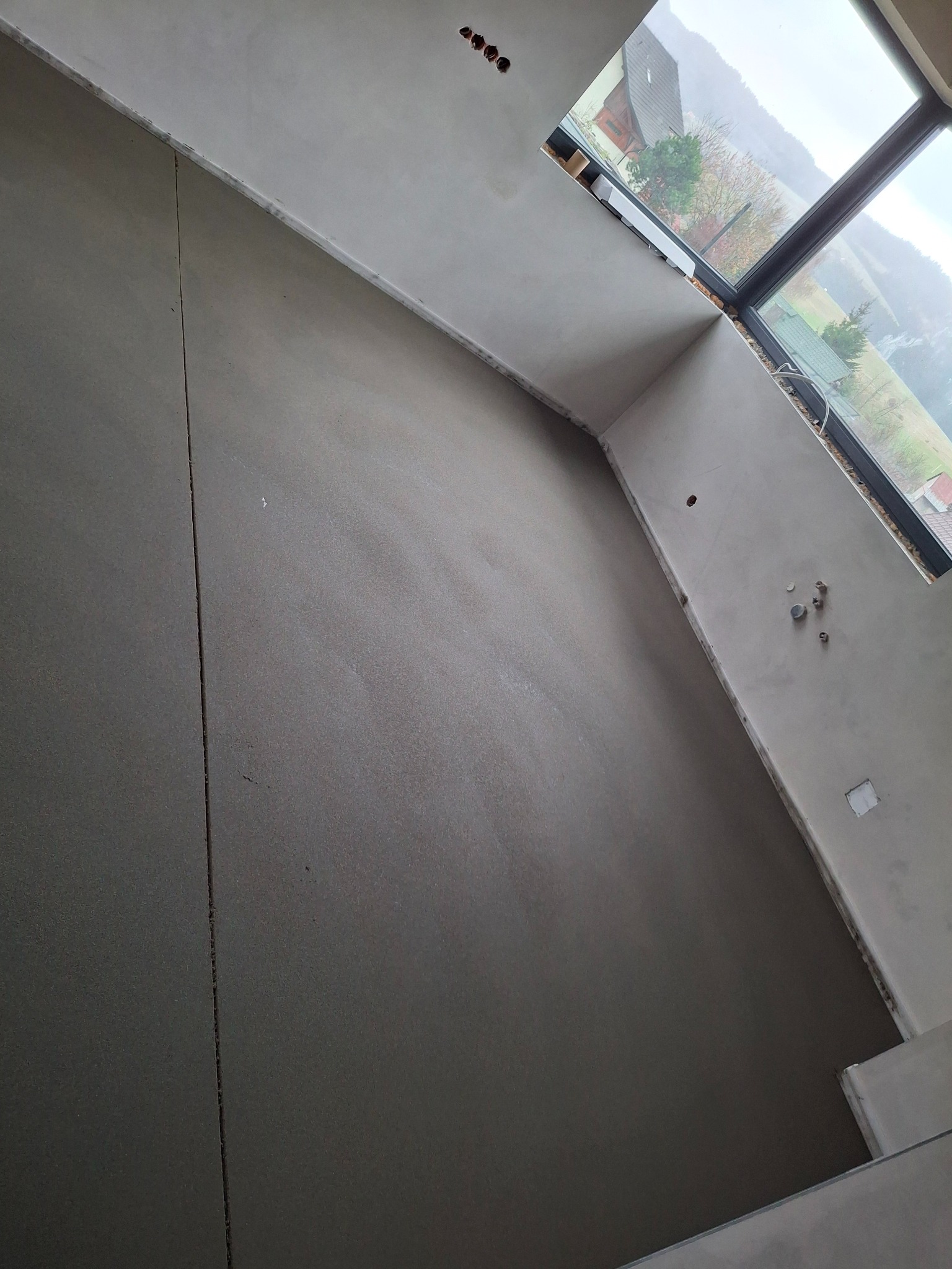 Świeżo wylana, gładka posadzka w jasnym wnętrzu z dużym oknem. Widoczne dylatacje w posadzce i fragment górskiego krajobrazu za oknem.