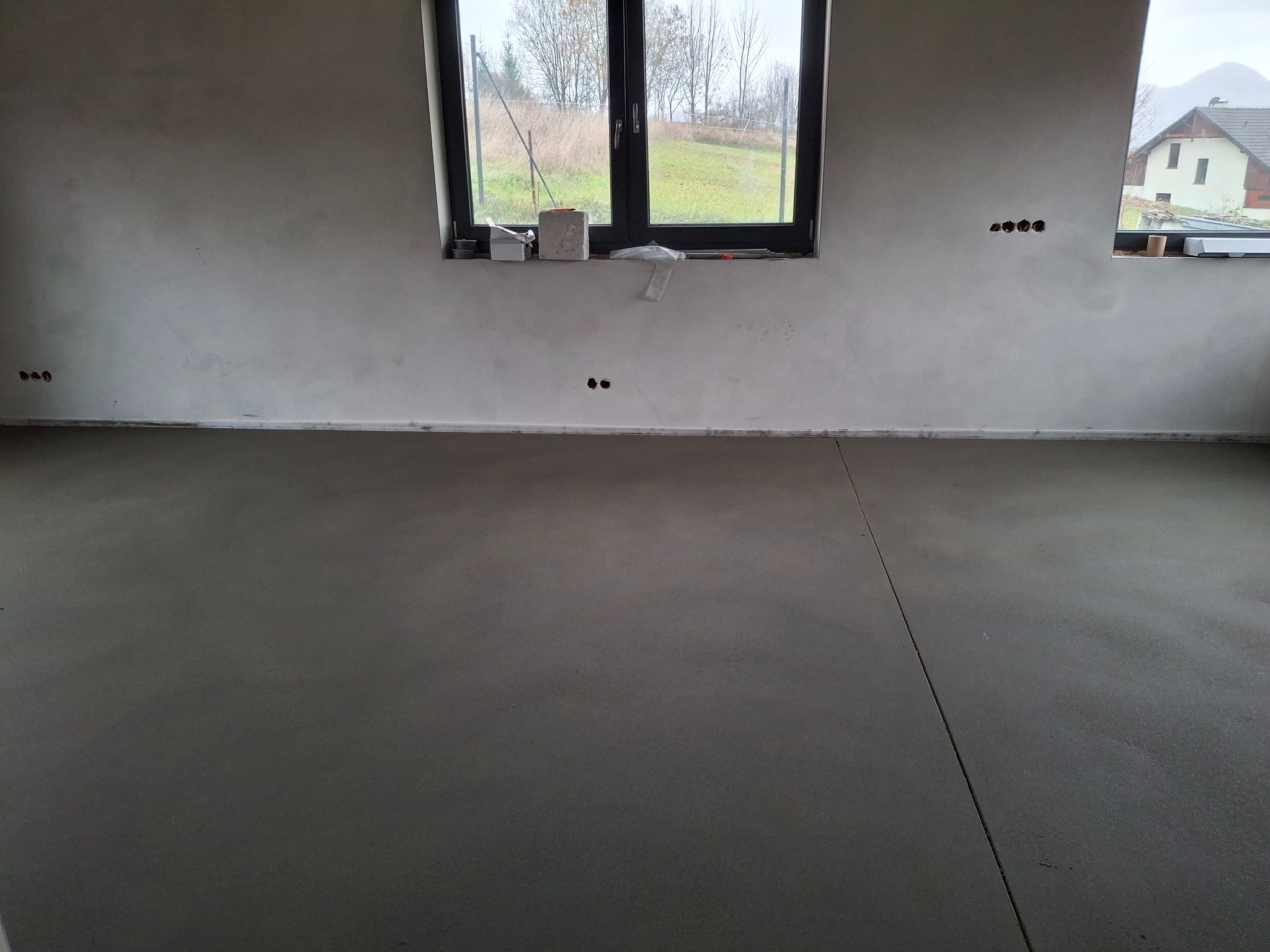 Świeżo wylana, szara posadzka w surowym wnętrzu. Widoczne okna z widokiem na zewnątrz i przygotowane otwory na gniazdka elektryczne w ścianie.