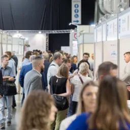 Tłum ludzi na targach branżowych, widoczne stoiska z logo firm i produkty. Tabliczka z napisem 'Toilets' w tle.