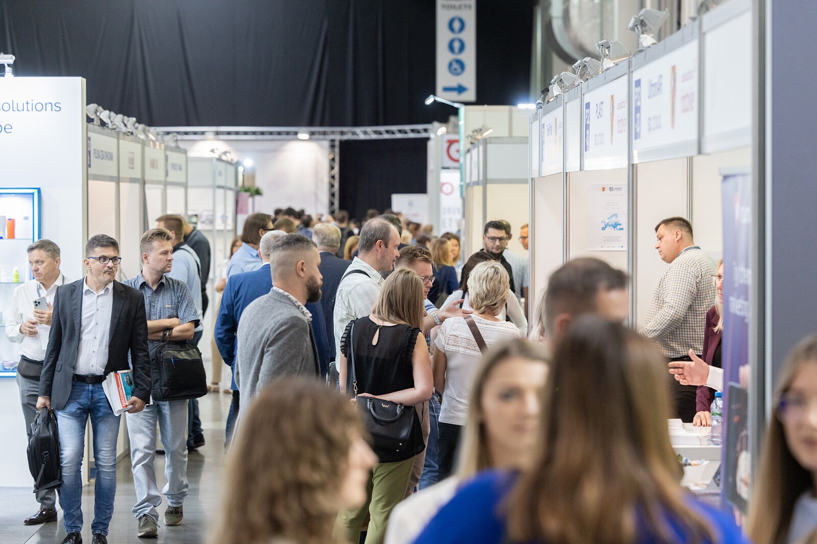 Tłum ludzi na targach branżowych, widoczne stoiska z logo firm i produkty. Tabliczka z napisem 'Toilets' w tle.