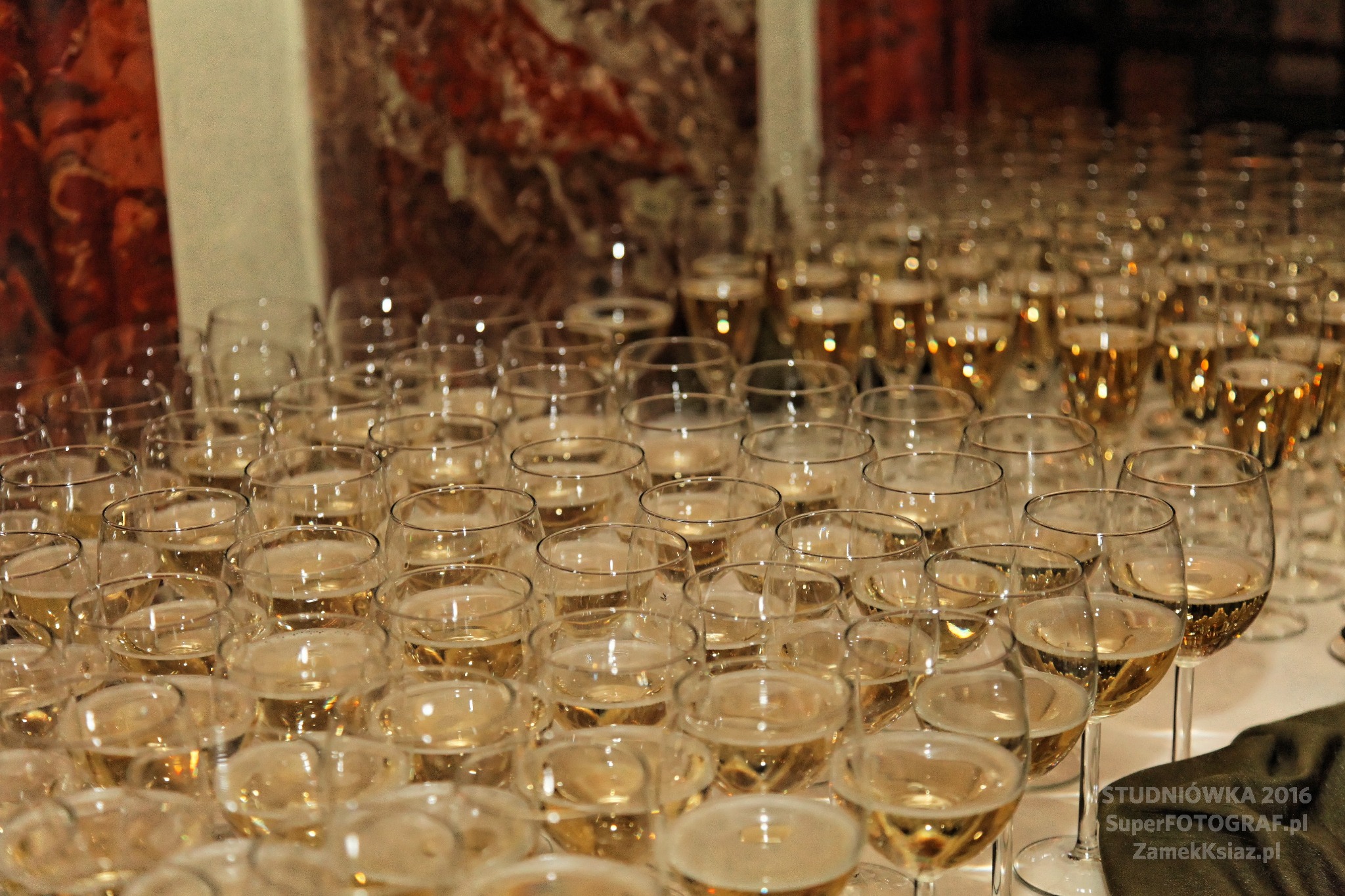 Ustawione w rzędach kieliszki wypełnione szampanem na stole bankietowym, przygotowane na imprezę. W tle marmurowa ściana i delikatne oświetlenie.