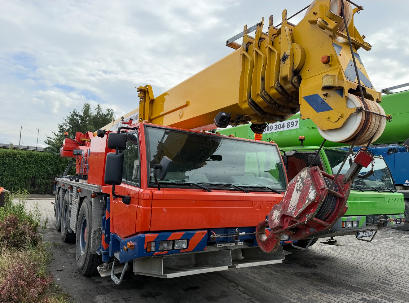 Czerwono-żółty dźwig Fauna na placu, z widocznym hakiem i fragmentami innych pojazdów w tle. Dźwig gotowy do pracy, widok z dołu podkreśla jego wielkość.