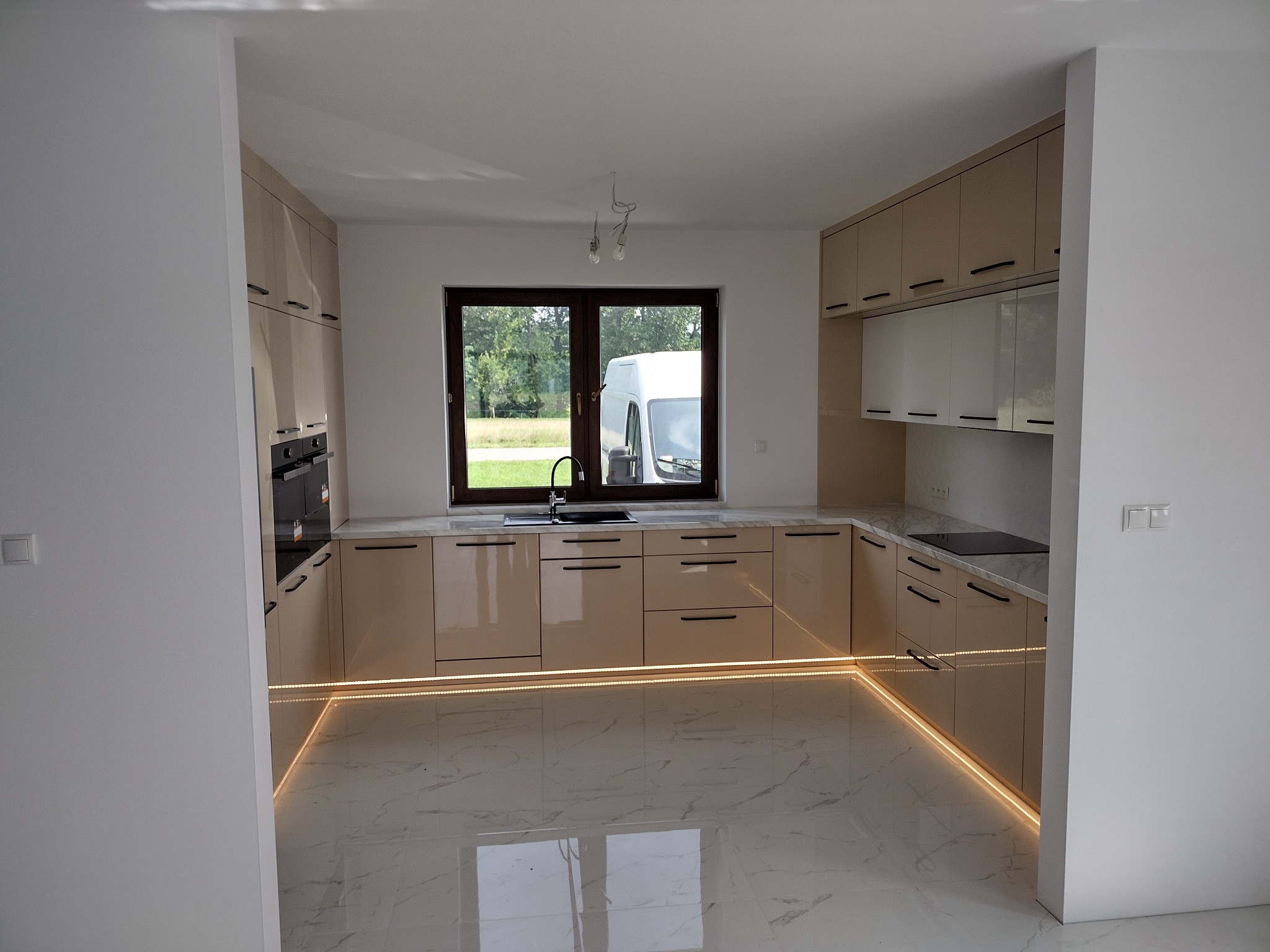 Beżowa kuchnia z oświetleniem LED przy podłodze, czarne, długie uchwyty, marmurowe płytki podłogowe, okno z widokiem na zieleń.