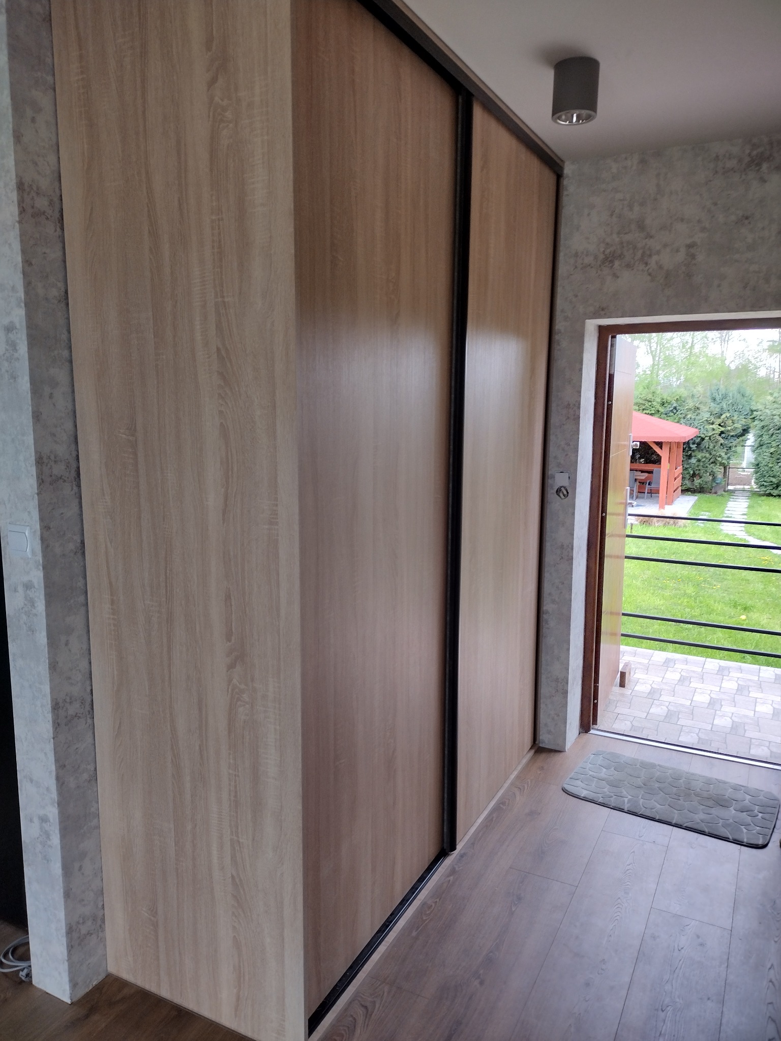Jasna szafa przesuwna z drewnianymi drzwiami na tle wejścia do domu z widokiem na ogród i altanę. Nowoczesny design, minimalistyczny styl.