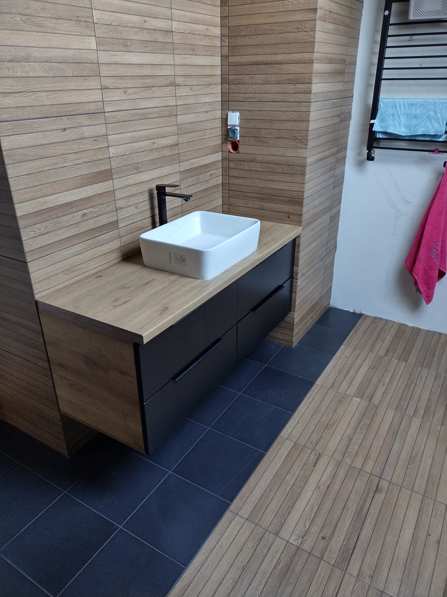 Nowoczesna łazienka z drewnopodobnymi płytkami ściennymi i podłogowymi. Umywalka nablatowa na szafce z czarnymi szufladami. Minimalistyczna armatura.