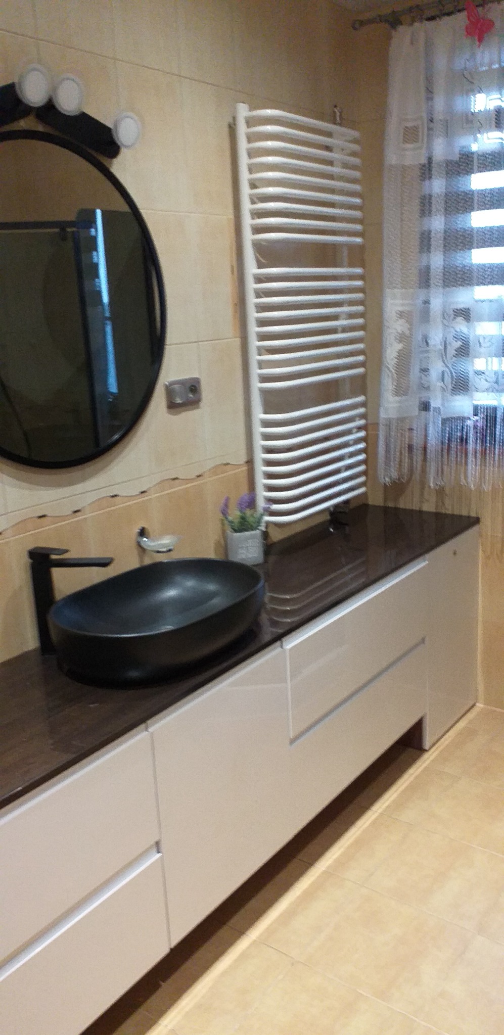 Nowoczesna łazienka z czarną, owalną umywalką na szafce z jasnymi frontami, obok grzejnik drabinkowy i okrągłe lustro z oświetleniem punktowym.