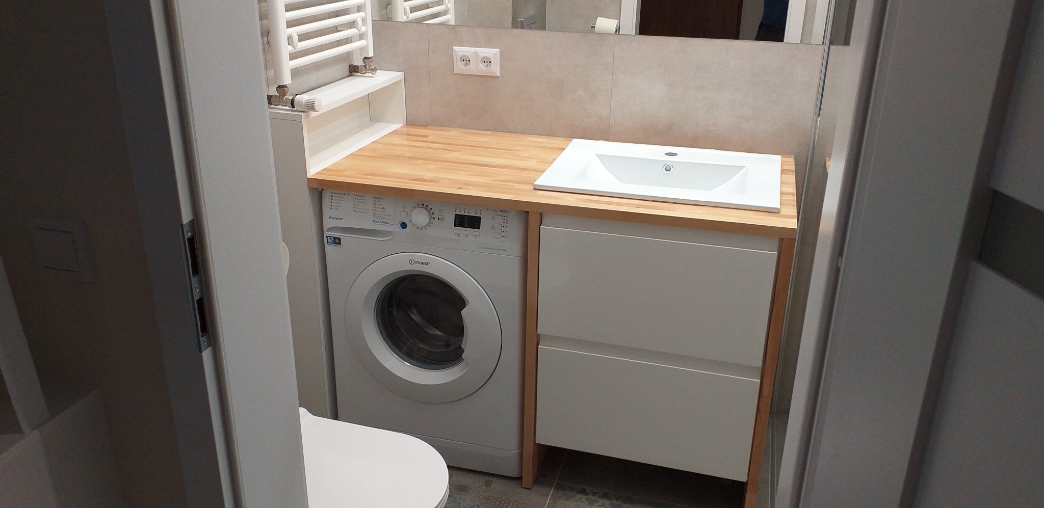 Funkcjonalna łazienka z pralką zabudowaną pod drewnianym blatem z umywalką. Białe szafki z dwoma szufladami i widoczny fragment toalety.