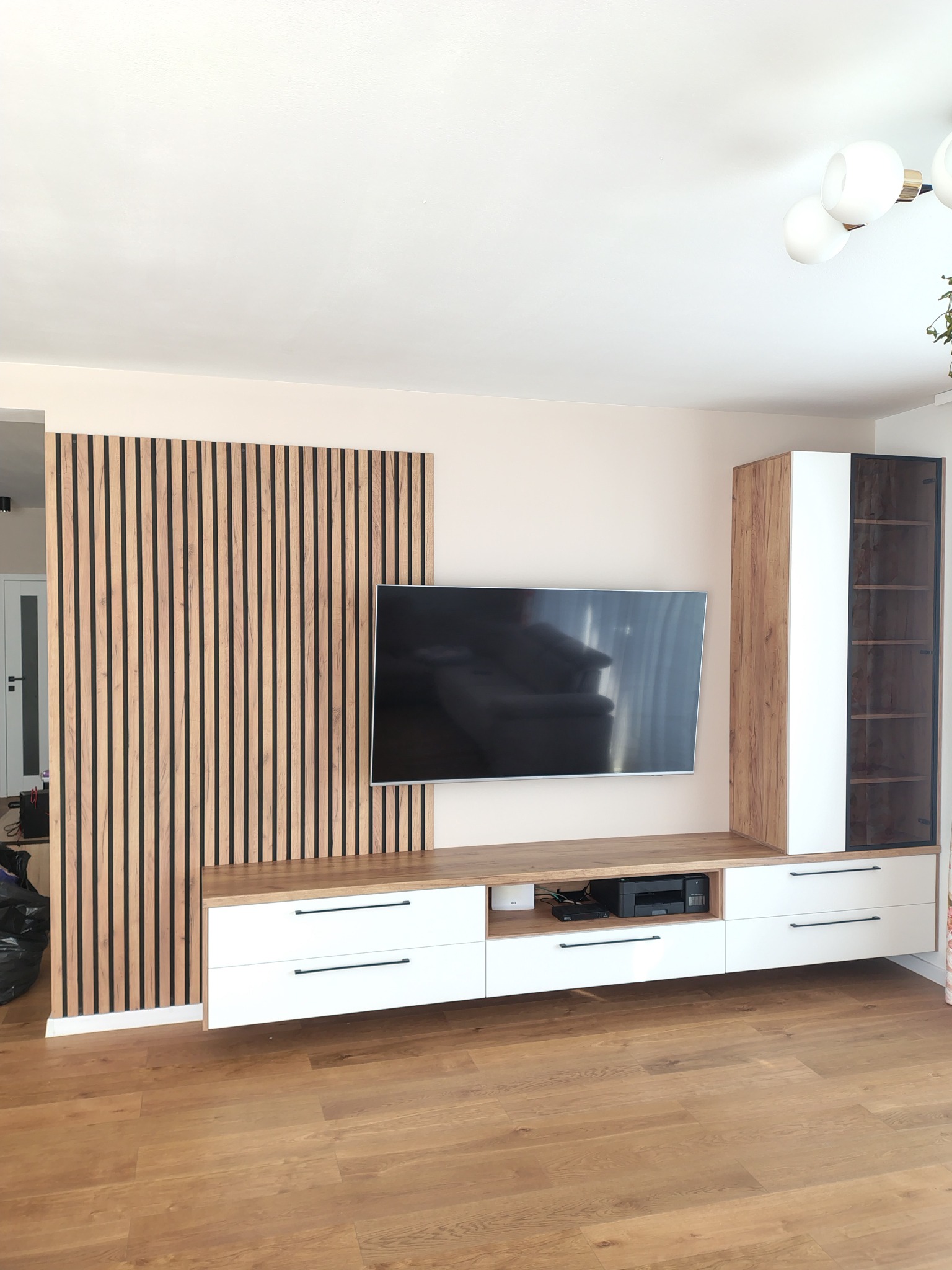 Nowoczesna zabudowa ściany TV z lamelami i szafkami w bieli i drewnie. Minimalistyczny design, drewniana podłoga, jasne wnętrze. Telewizor zawieszony na ścianie.