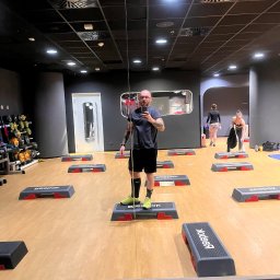 PIOTR WARUNEK TRENINGI - Mężczyzna robi selfie w lustrze sali fitness. W tle stepy Reebok, hantle i ćwiczące osoby. Oświetlenie sufitowe, ciemne ściany. Ujęcie z dołu.