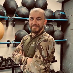 PIOTR WARUNEK TRENINGI - Uśmiechnięty mężczyzna w sportowej odzieży moro, skrzyżowane ręce na tle ściany z piłkami lekarskimi i hantlami w siłowni.