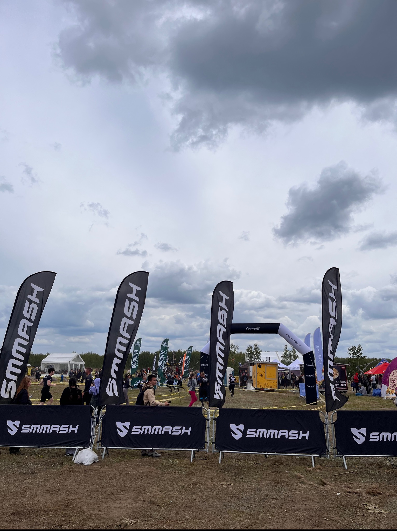 Zawody sportowe na zewnątrz z bramą OstroVit, flagami SMMASH i uczestnikami w tle pod pochmurnym niebem. Event sportowy.