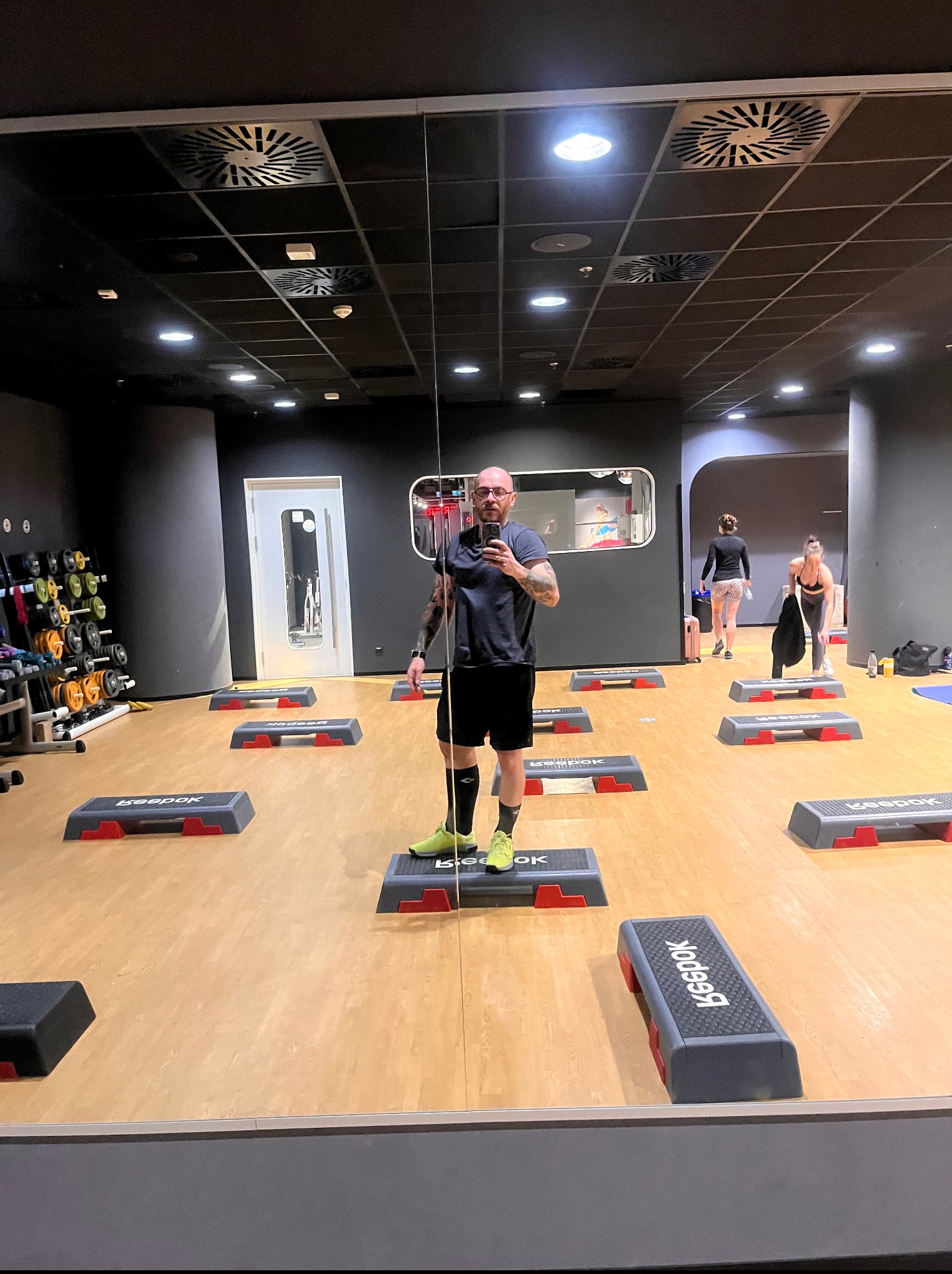 Mężczyzna robi selfie w lustrze sali fitness. W tle stepy Reebok, hantle i ćwiczące osoby. Oświetlenie sufitowe, ciemne ściany. Ujęcie z dołu.