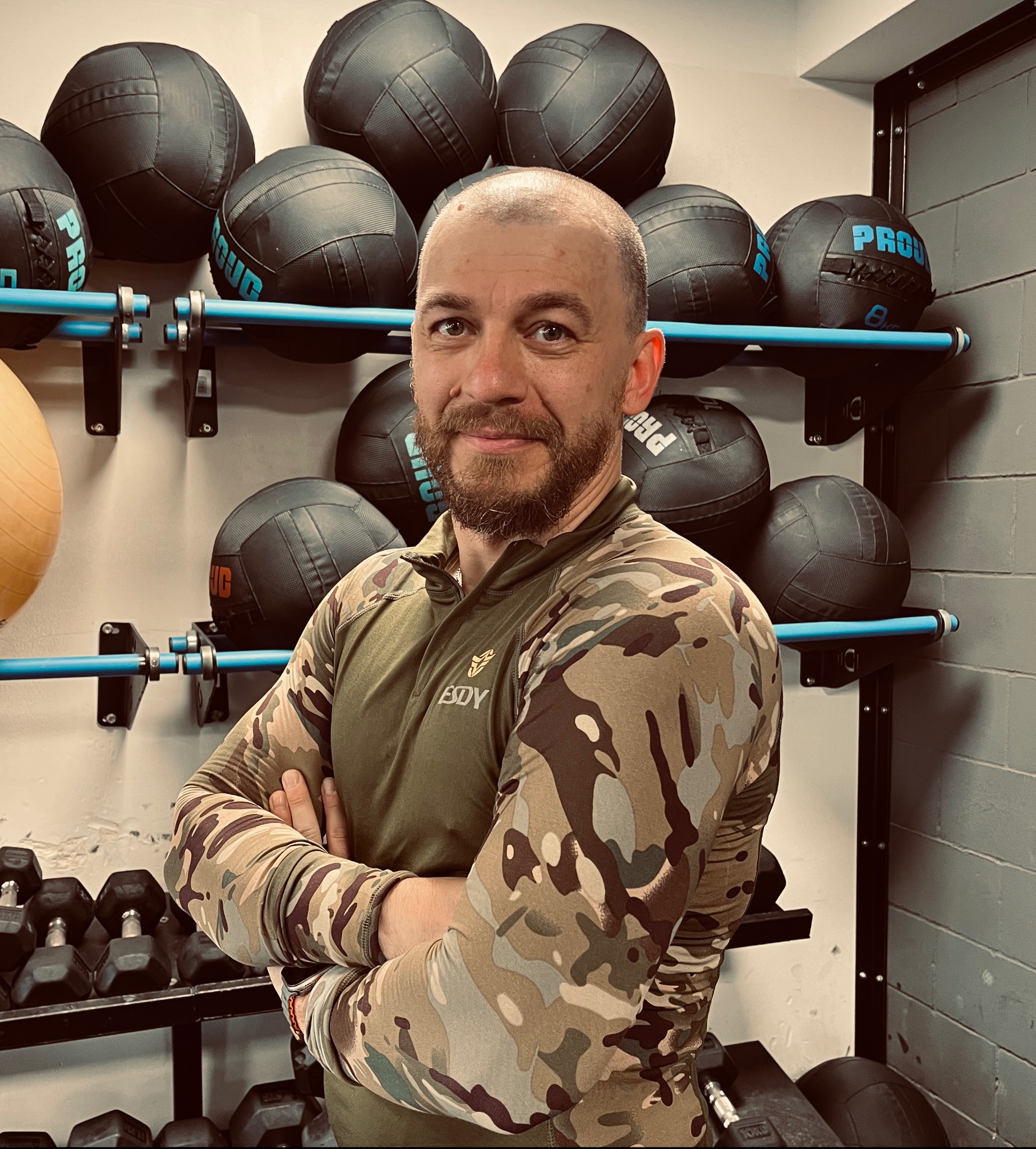 Uśmiechnięty mężczyzna w sportowej odzieży moro, skrzyżowane ręce na tle ściany z piłkami lekarskimi i hantlami w siłowni.