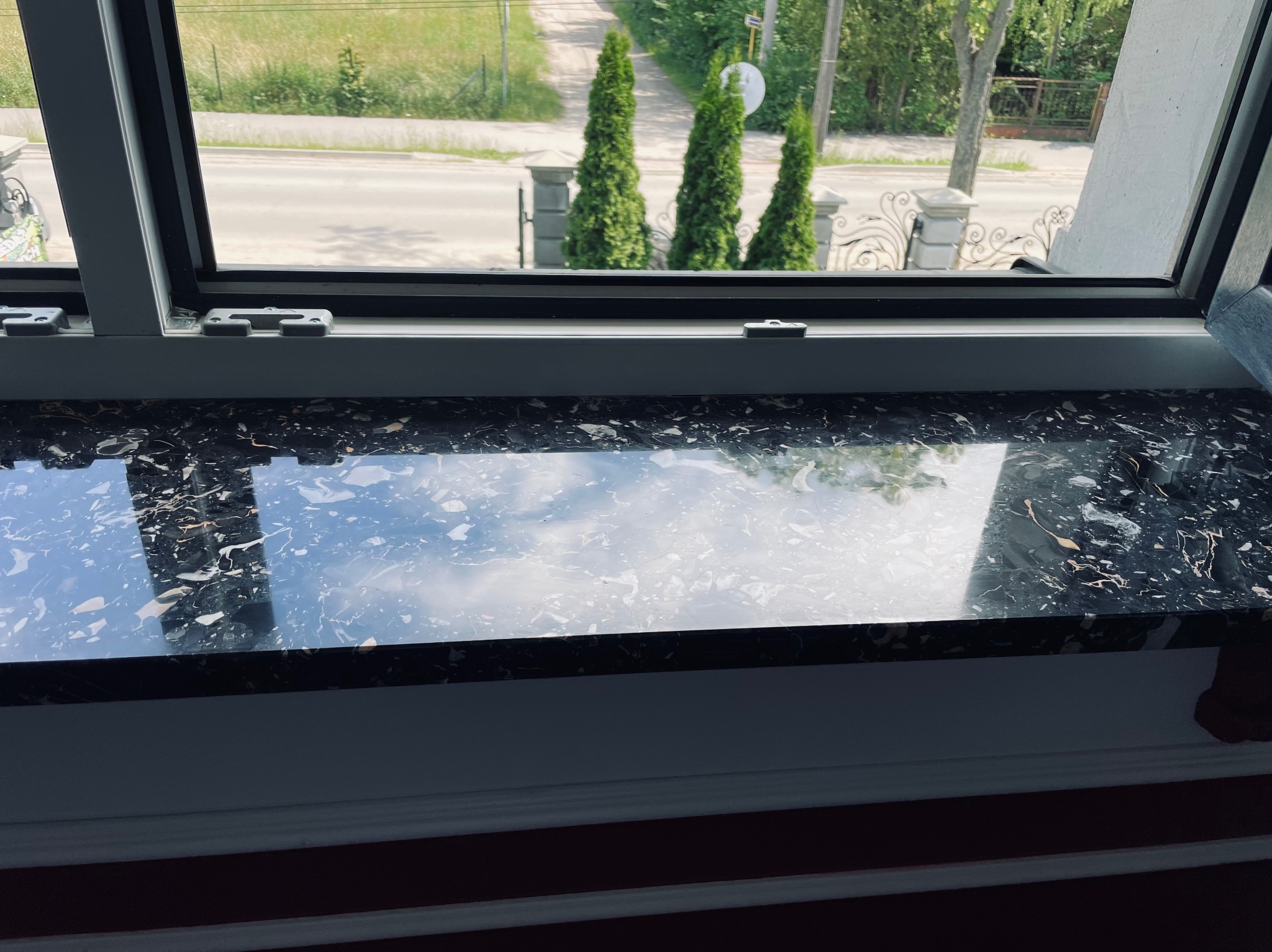 Czarny, błyszczący parapet z kamienia w oknie, z widokiem na ulicę i ozdobne tuje. Delikatne odbicie nieba na powierzchni kamienia.