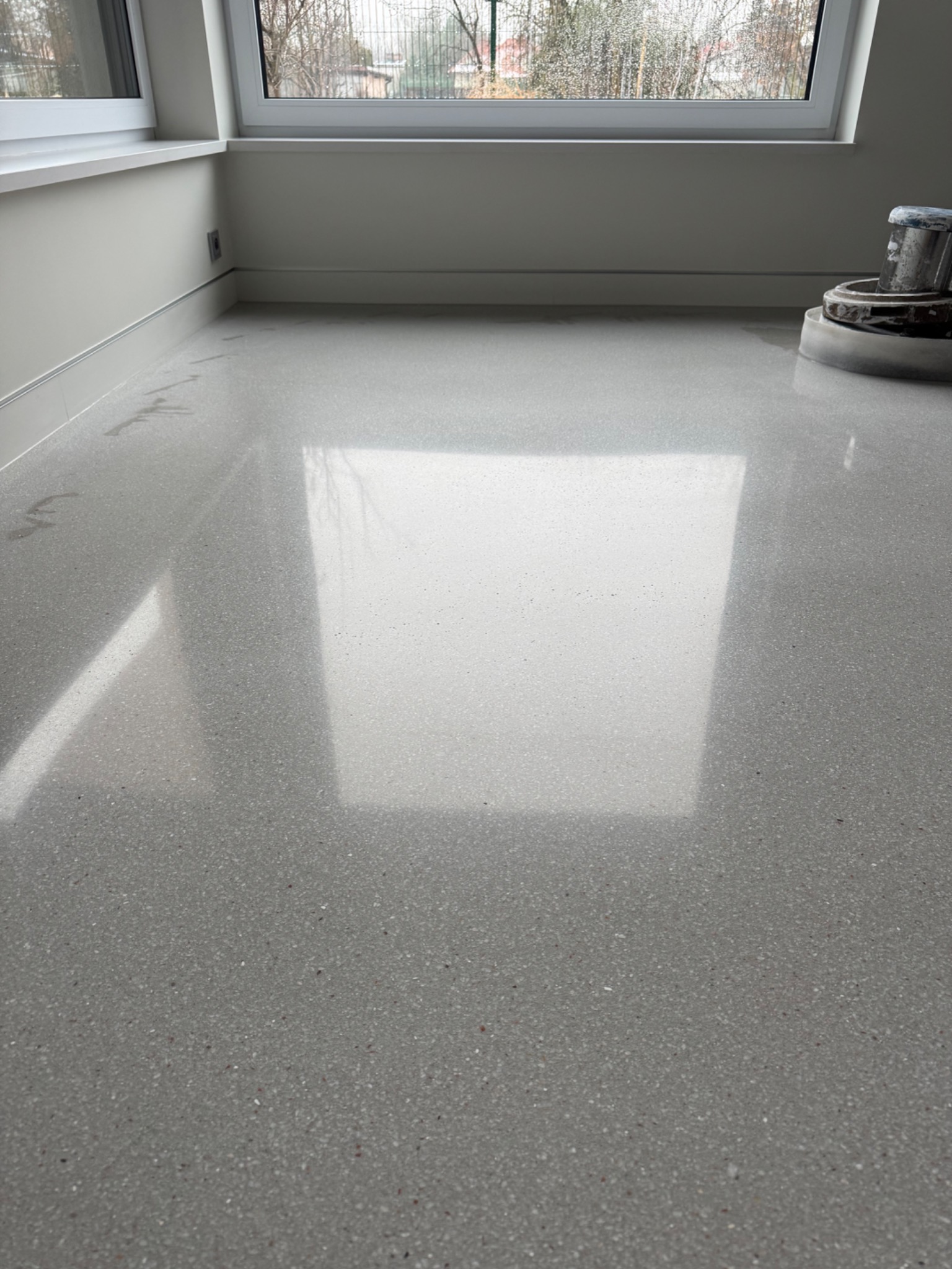 Szlifowana, jasnoszara posadzka z lastryko w pokoju z oknem. Fragment maszyny do polerowania w prawym górnym rogu. Widoczne ślady wody na powierzchni.