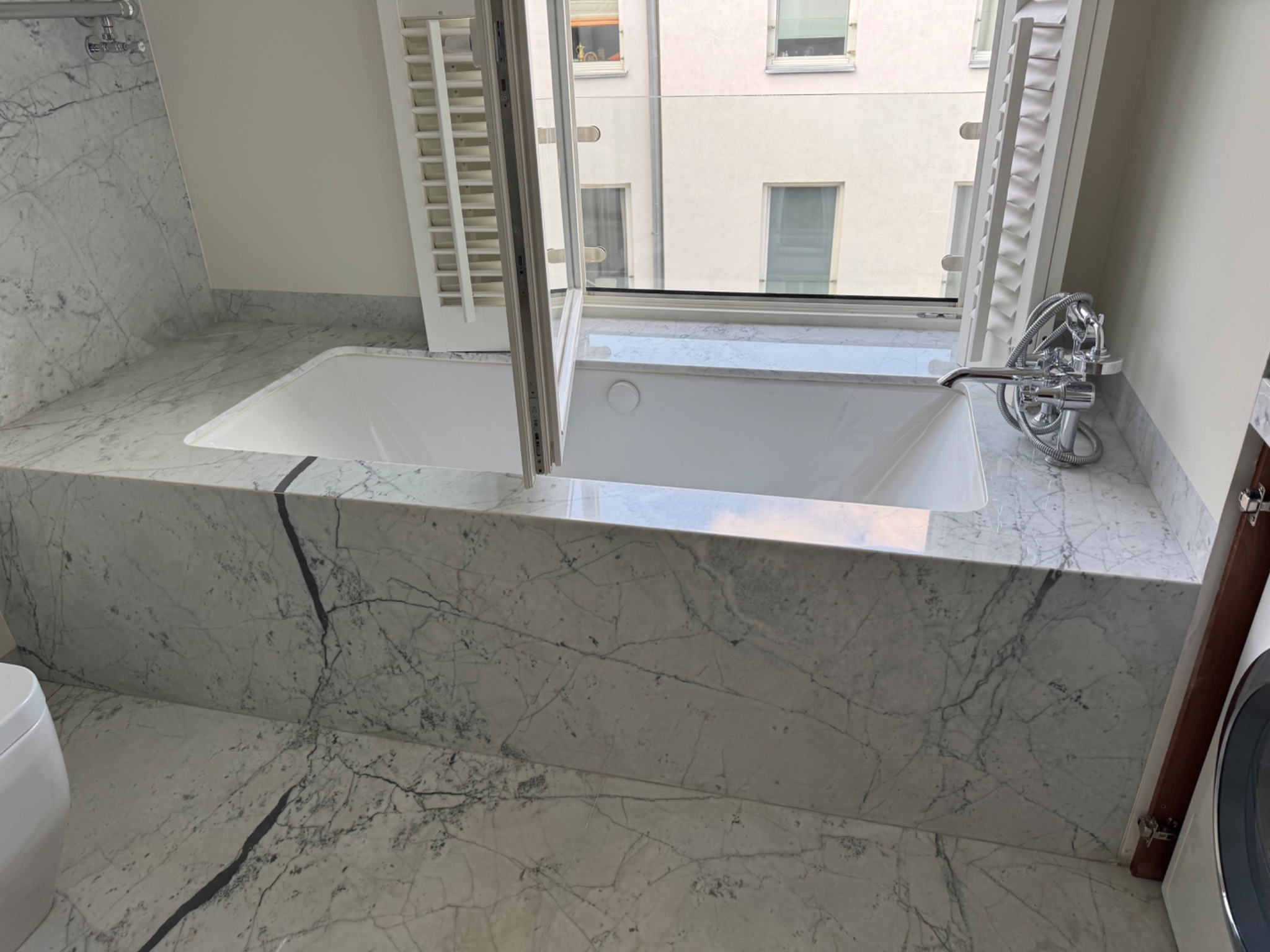 Elegancka wanna obudowana marmurem z wyraźnym żyłowaniem, okno z białymi okiennicami w tle. Bateria chromowana. Widok na budynek z okna.