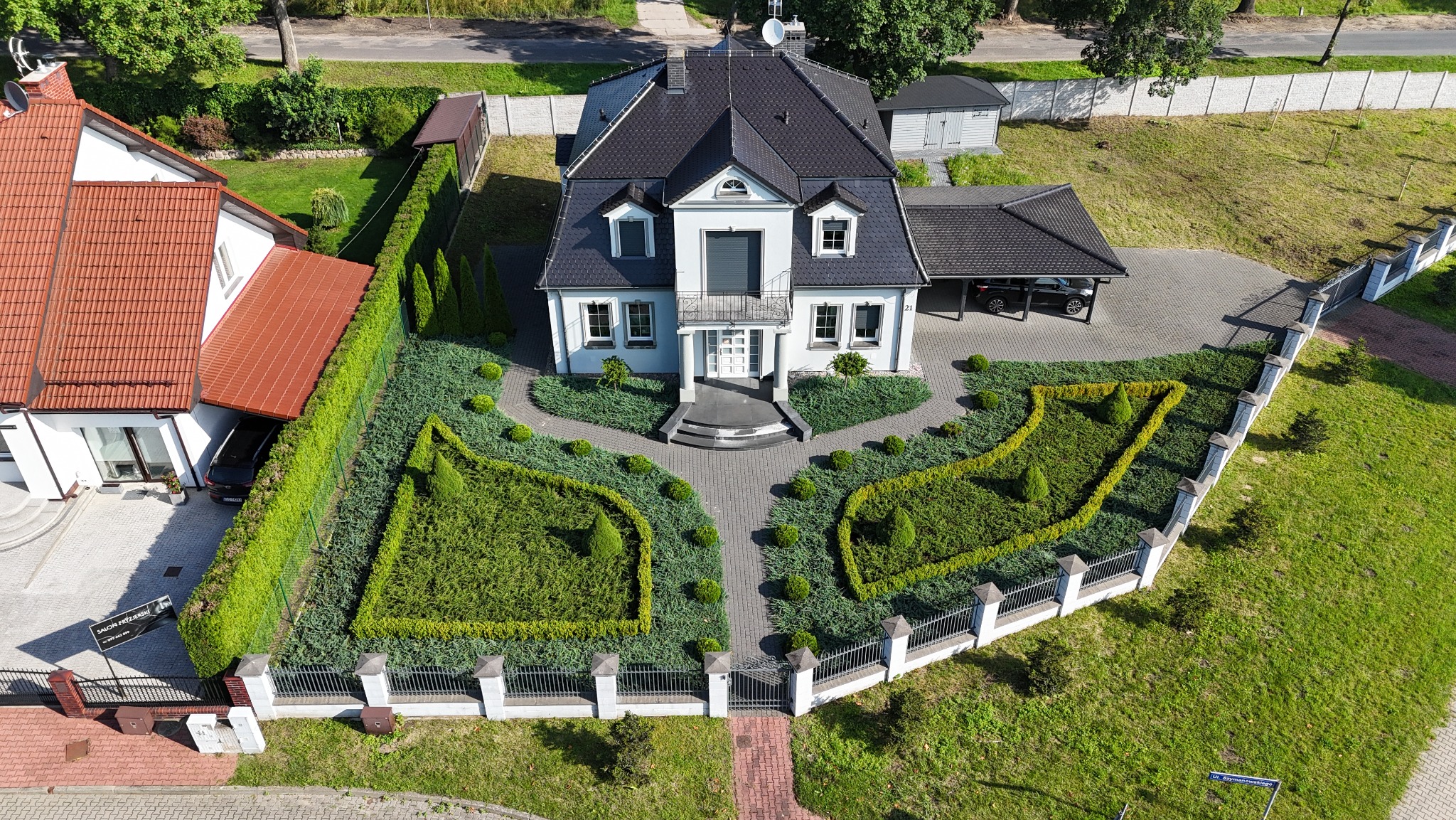 Widok z góry na dom z zadbanym ogrodem: geometryczne rabaty z iglakami, ścieżki z kostki, ogrodzenie z białymi słupkami. Zadbany trawnik, równo przycięty żywopłot.